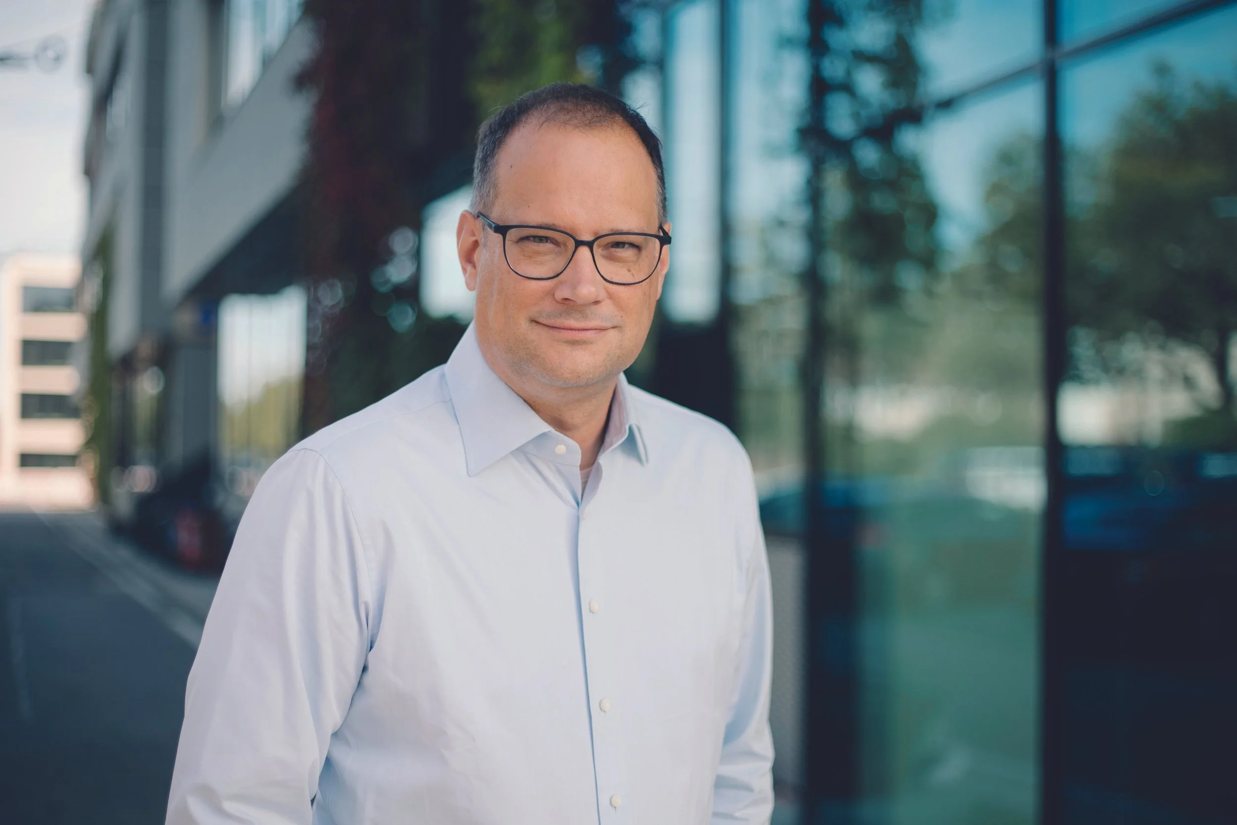 A professional headshot of a man in a dark suit, white shirt, and dark tie, smiling against a plain gray background.