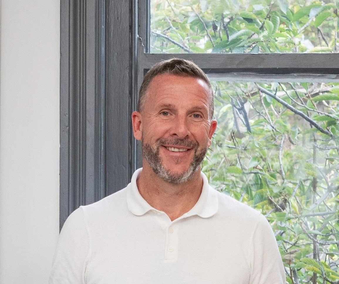 A smiling man with short hair and a beard, wearing a white polo shirt, standing indoors near a window with greenery outside.