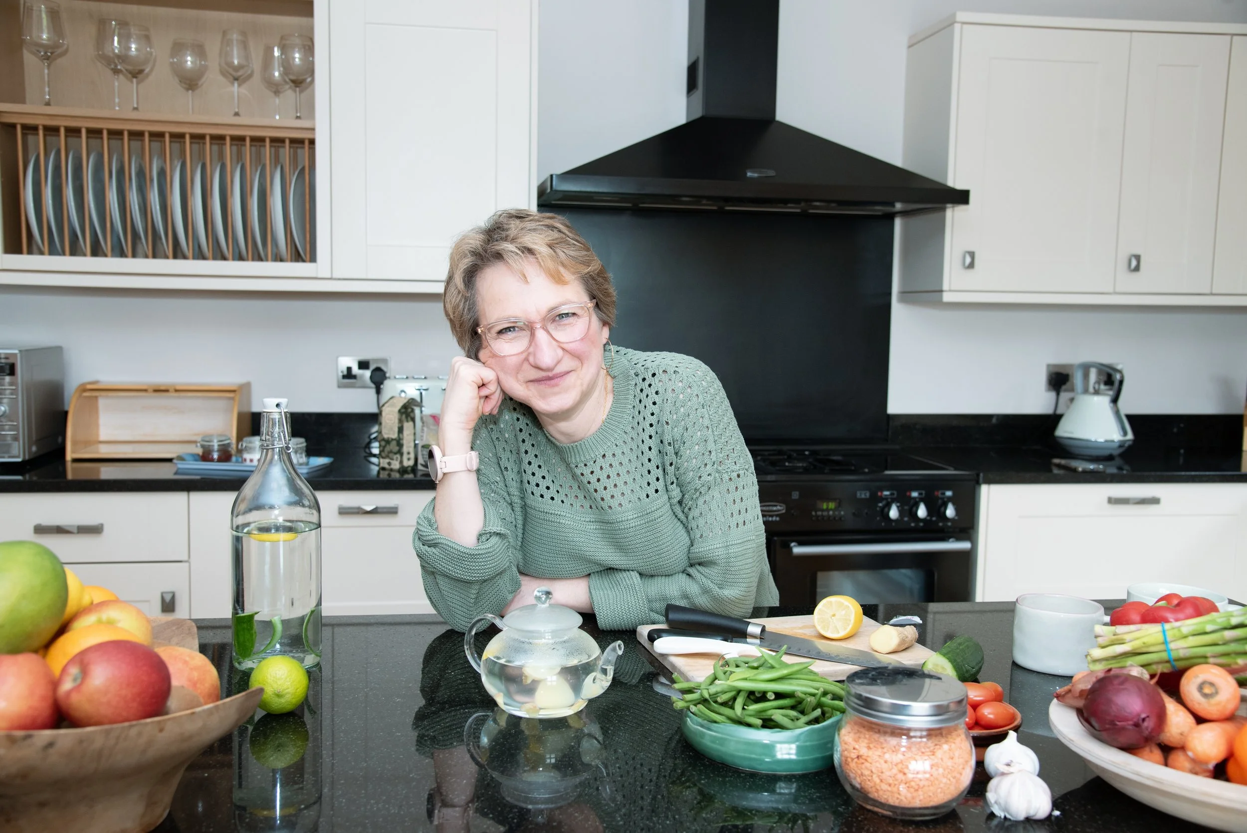 Sandra Dumas smiling in a bright, modern kitchen, leaning on a dark granite countertop surrounded by fresh fruits, vegetables, lentils, and a glass teapot.