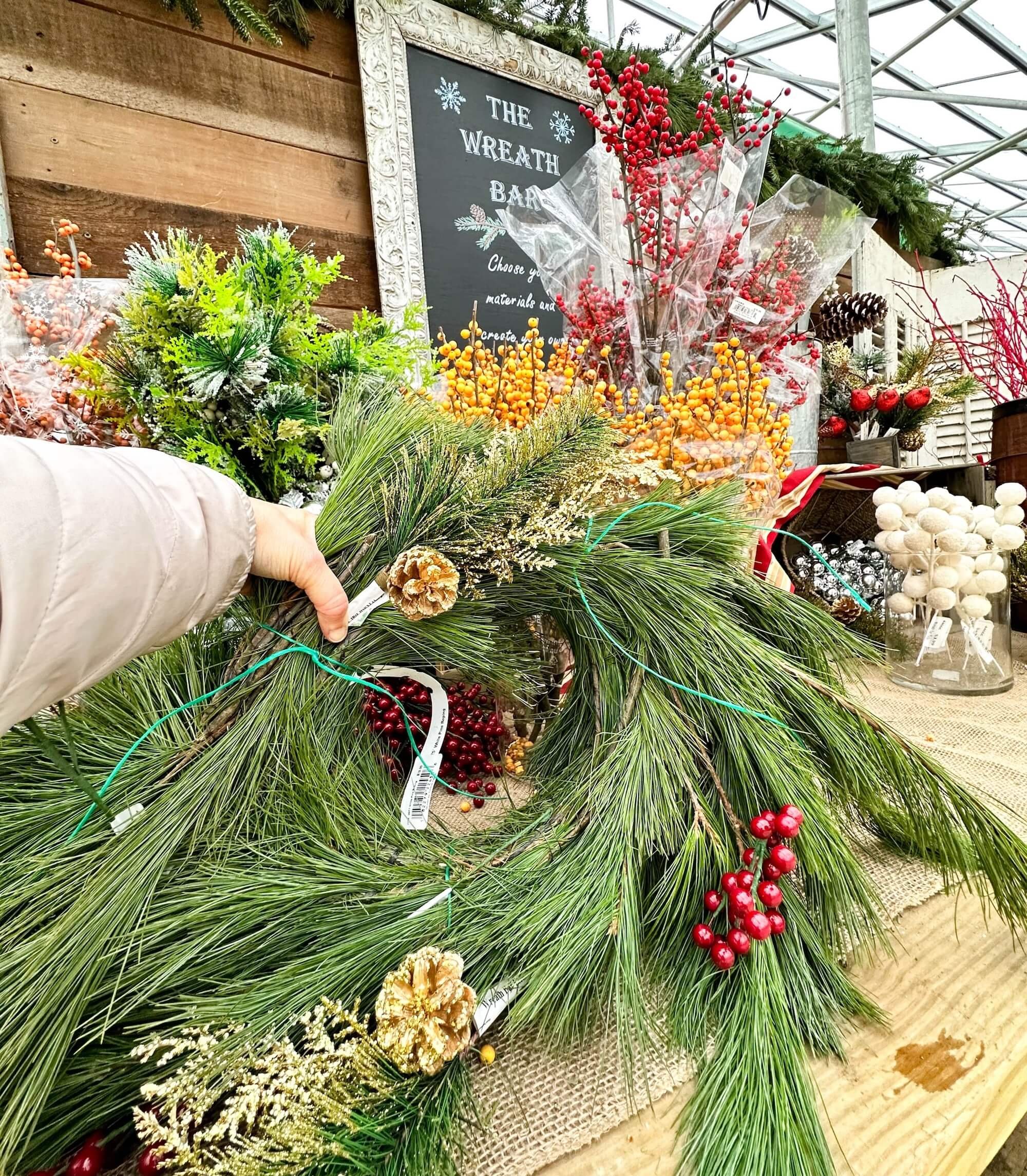 A hand holds a pine wreath decorated with red berries and pinecones at Timbuk Farms’ wreath bar, surrounded by holiday greenery and decorations.