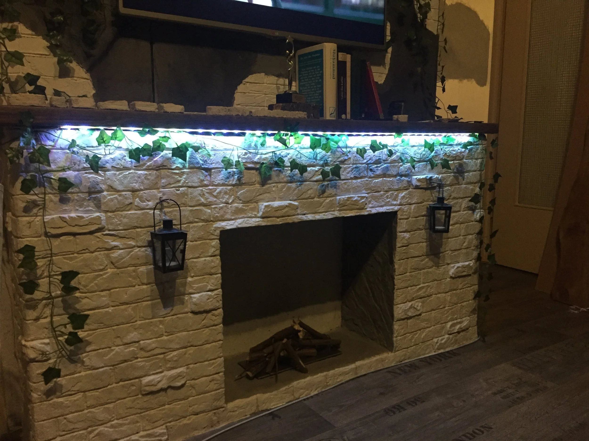 Stein Kamin mit Grünpflanzen und LED-Leiste, zwei Laternen, Bücher auf der oberen Ablage, Holzstücken im Feuerkasten, in einem Raum ohne Kaminabzug.