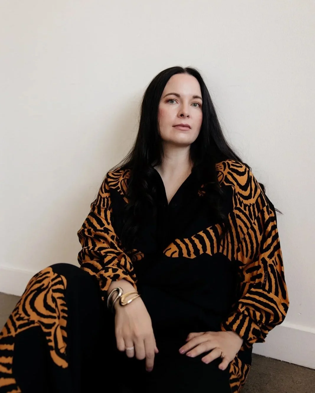 Sara Moon sitting against a plain white wall, wearing a black and orange tiger-patterned outfit, with her hand resting on her knee, looking at the camera.