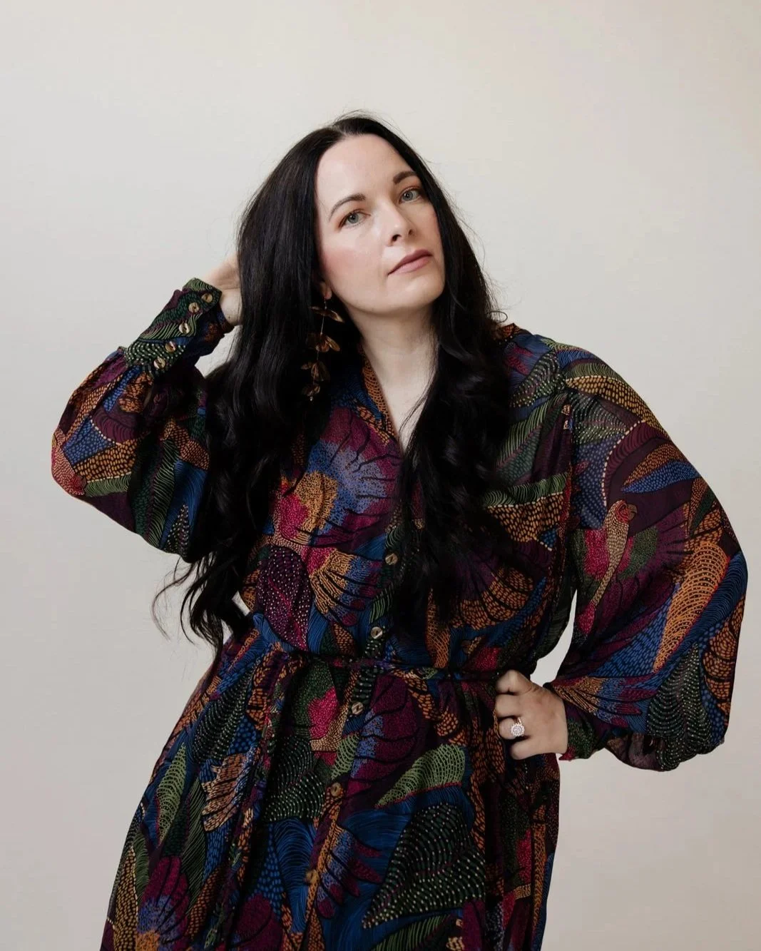 Sara Moon wearing a colorful patterned dress, posing with one hand on her hip and the other behind her head against a plain background.