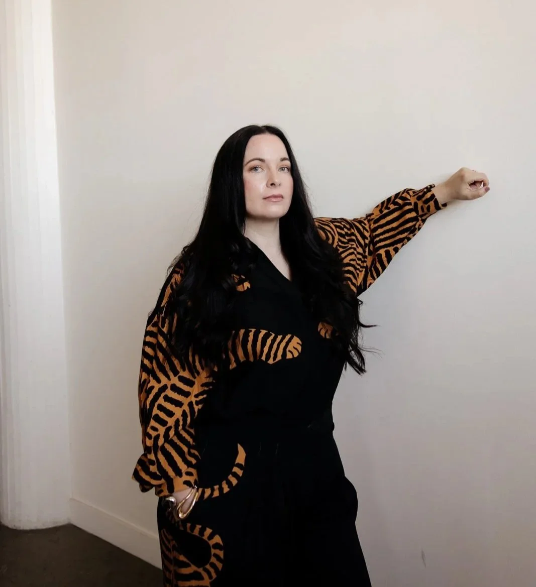 Sara Moon with long black hair standing against a plain white wall, wearing a black and orange tiger-stripe patterned shirt and black pants, with her arm extended to the side.