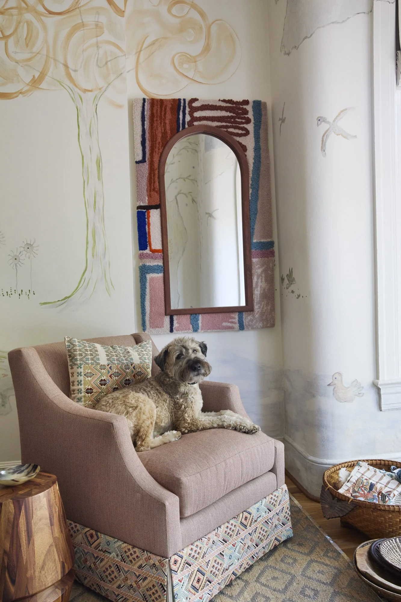 Pink upholstered chair with embroidered pillows beneath a colorful textile-framed mirror in the RBI studio.