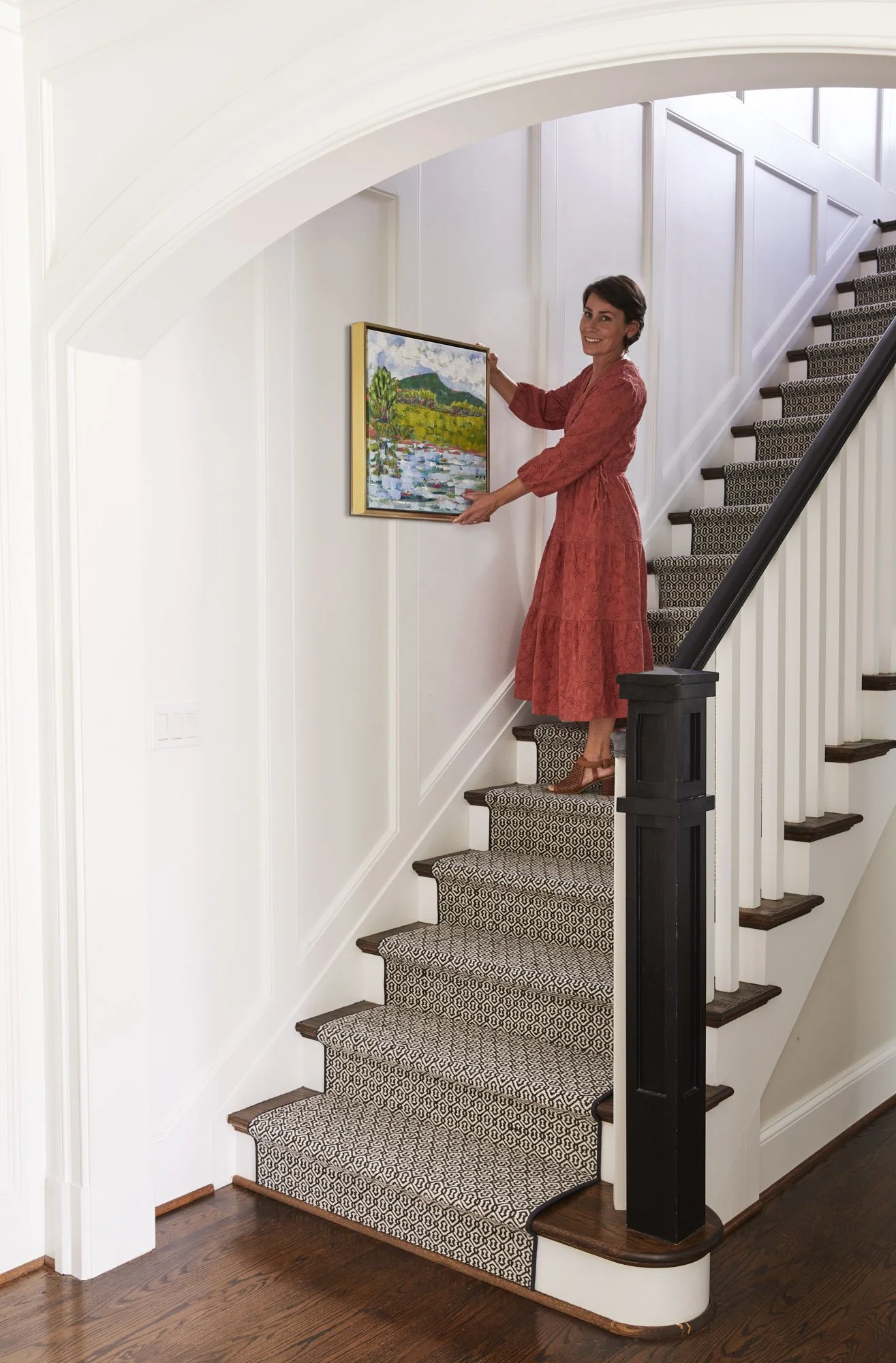 Regan in a red dress hanging framed artwork along a curved staircase with a patterned runner.