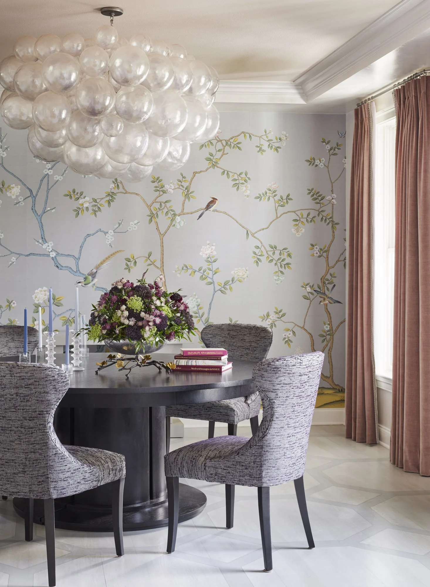 Round dining table with upholstered chairs, a clustered pendant light, floral bird wallpaper, flowers, and stacked books.