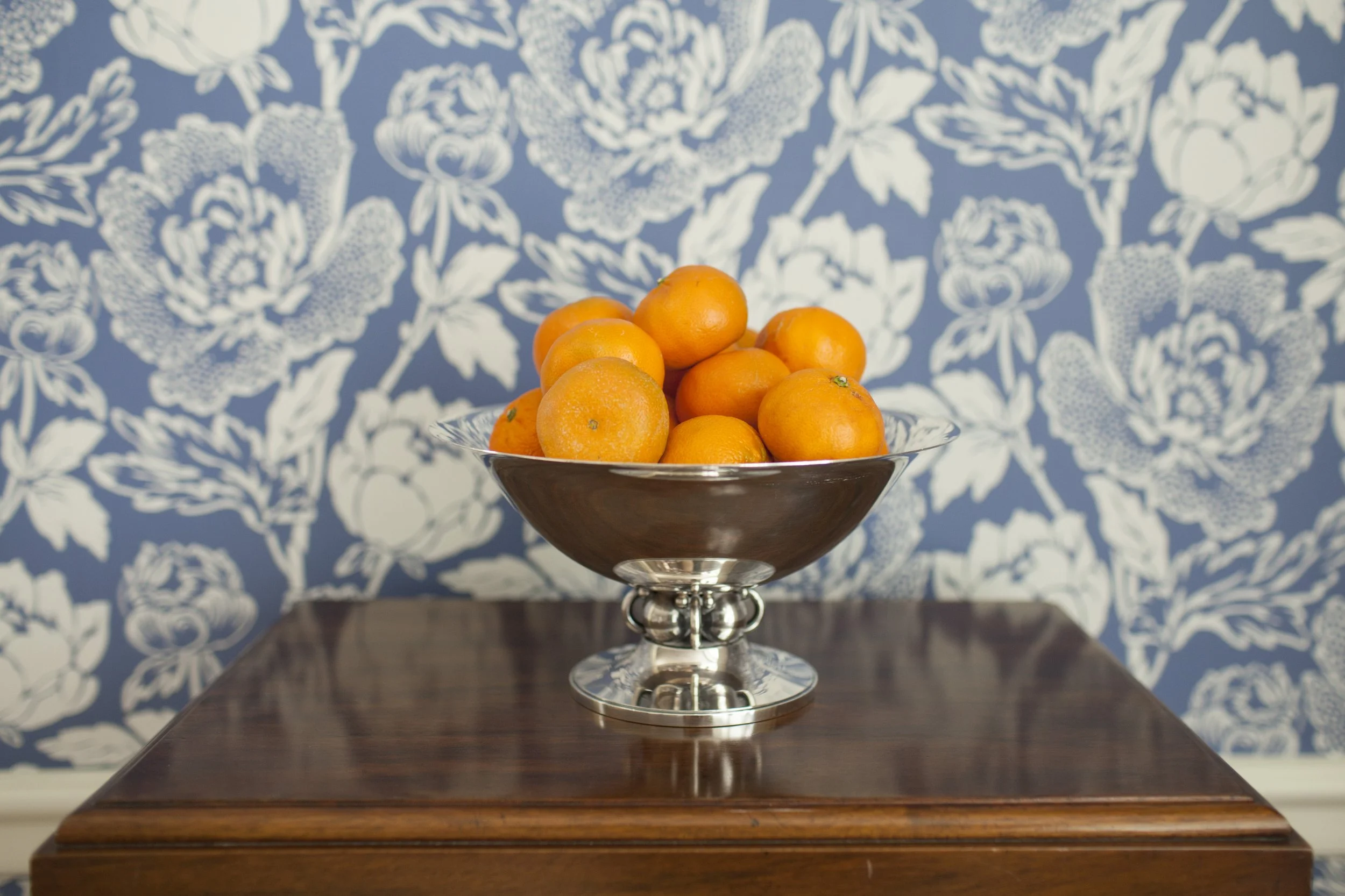 Silver bowl filled with oranges on a wood dining table against blue floral wallpaper.
