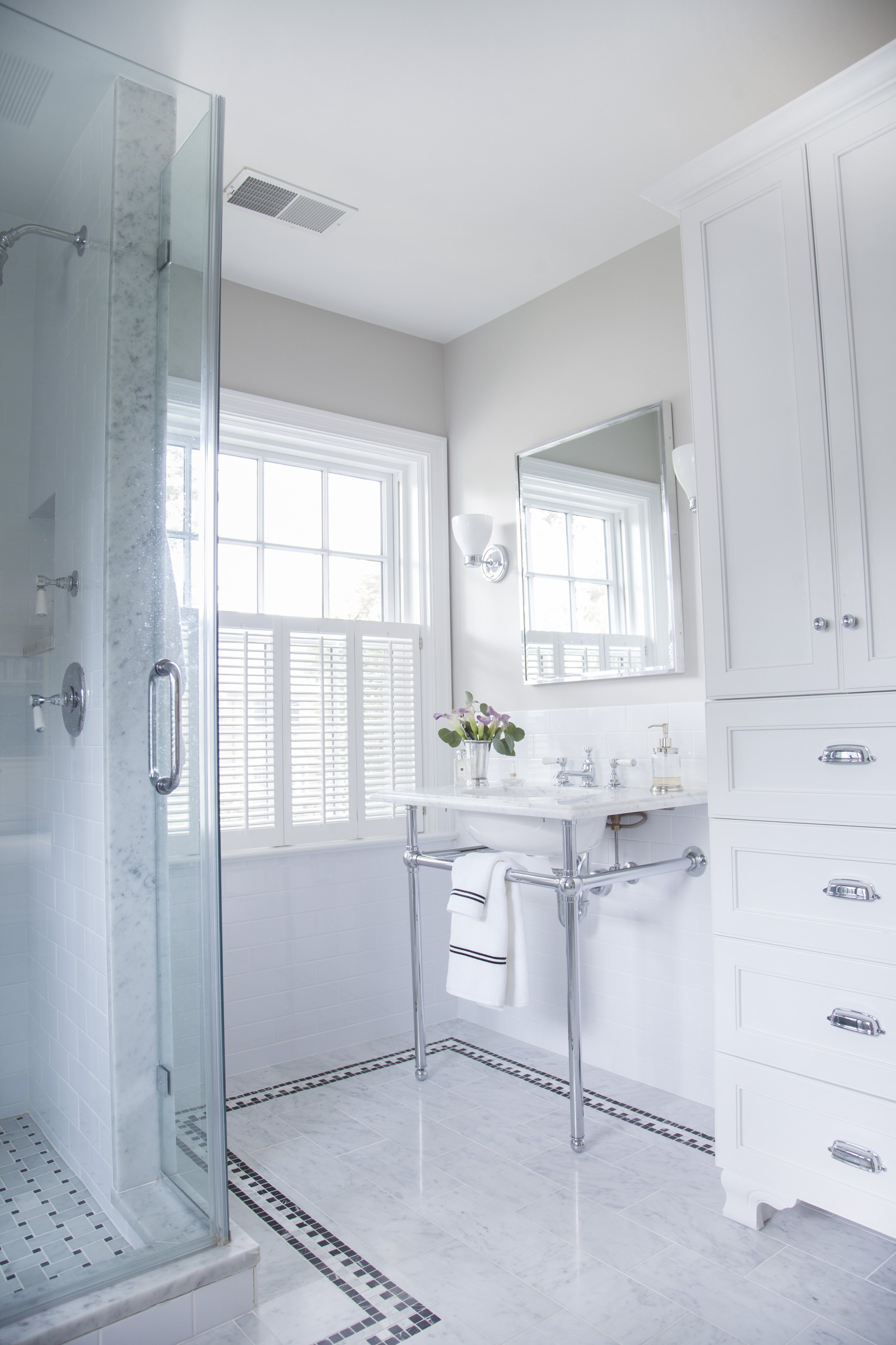 Light-filled bathroom with a marble shower, white vanity, large mirror, storage cabinet, and fresh flowers.