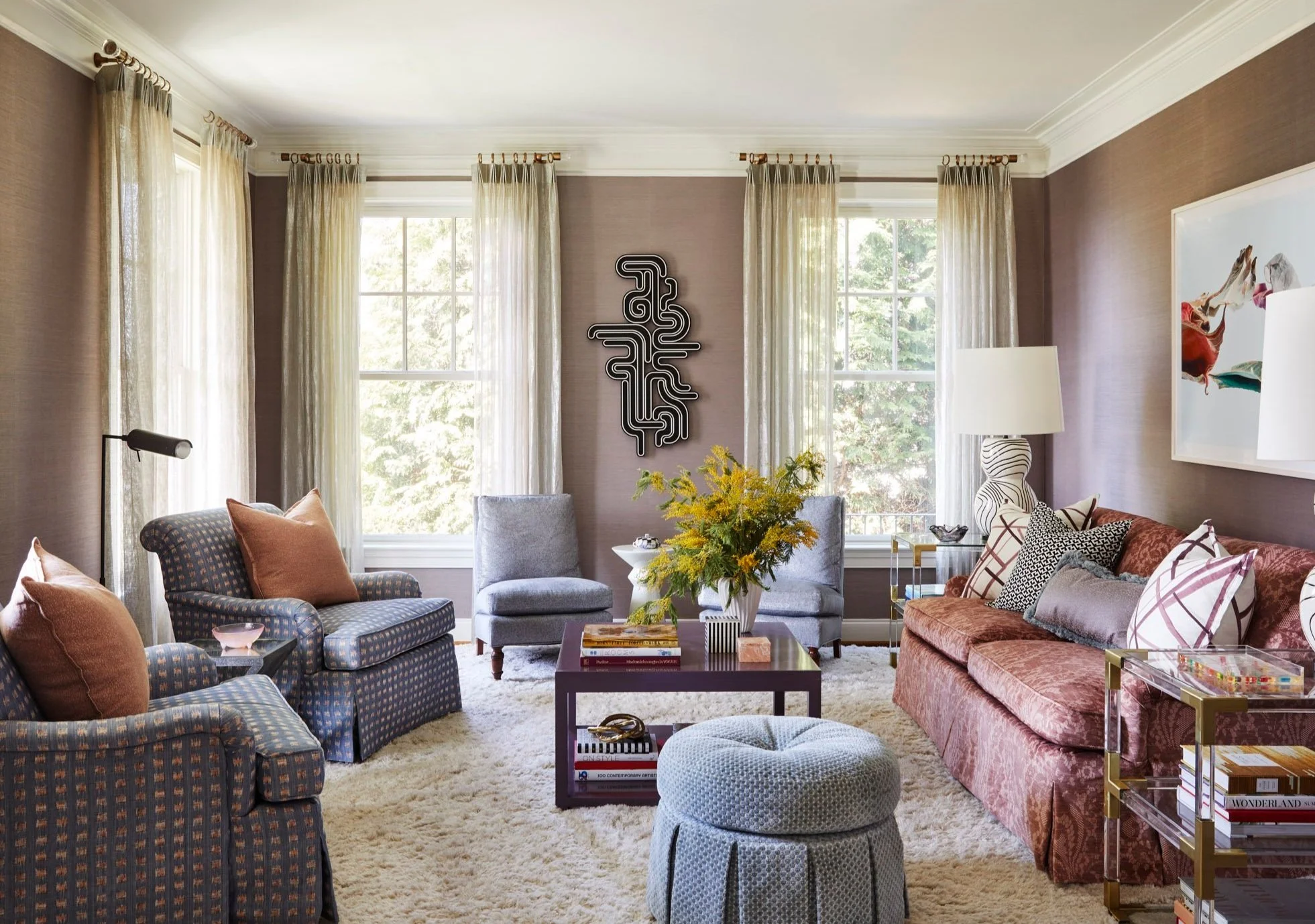 Living room with patterned armchairs, a warm brown sofa, layered pillows, abstract artwork, and large windows with sheer curtains.