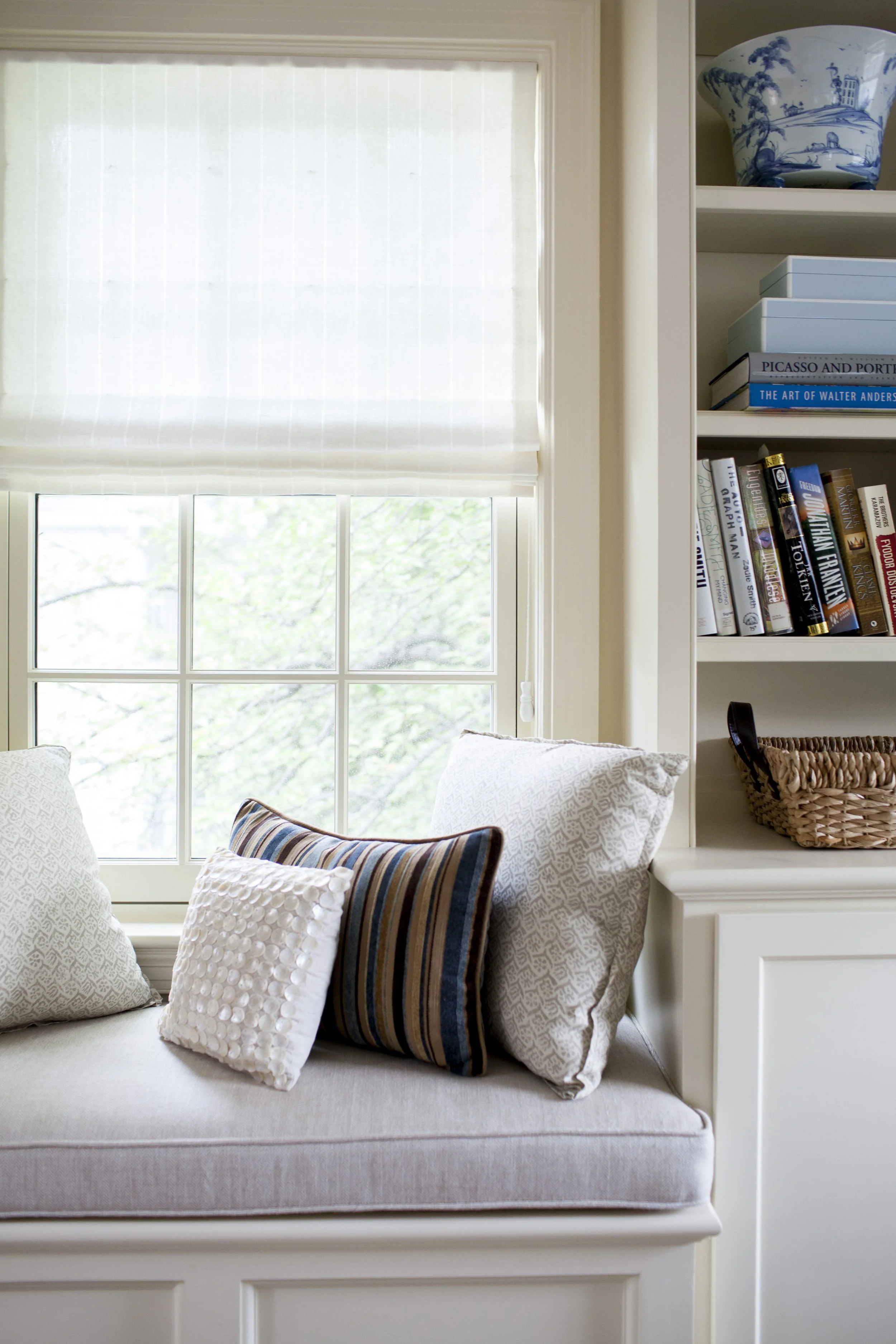 Cozy window seat with white blinds, patterned pillows, and built-in bookshelves filled with books and a woven basket.