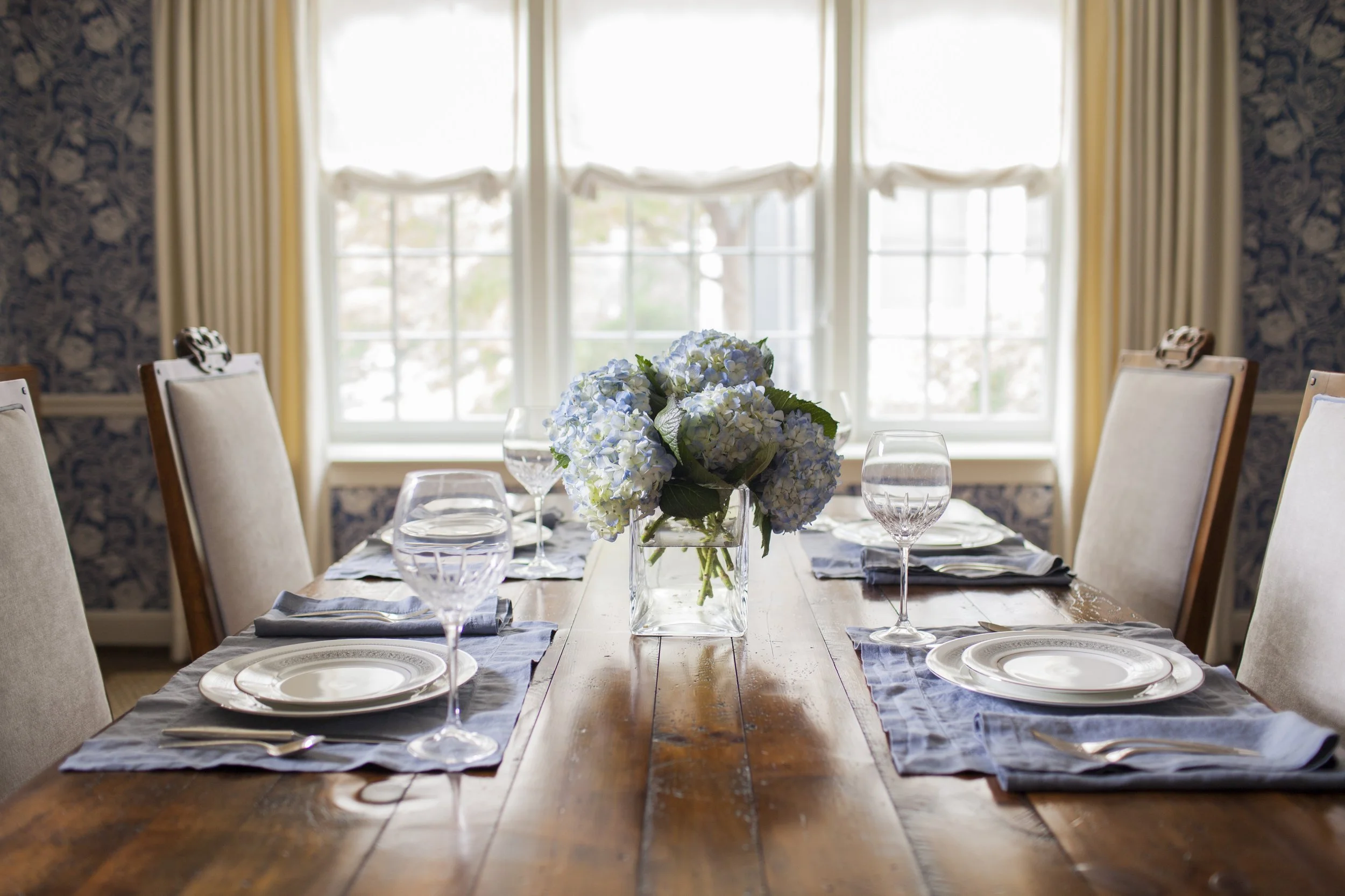 Formal dining table set with blue napkins, white plates, crystal glasses, and a centerpiece of blue hydrangeas.