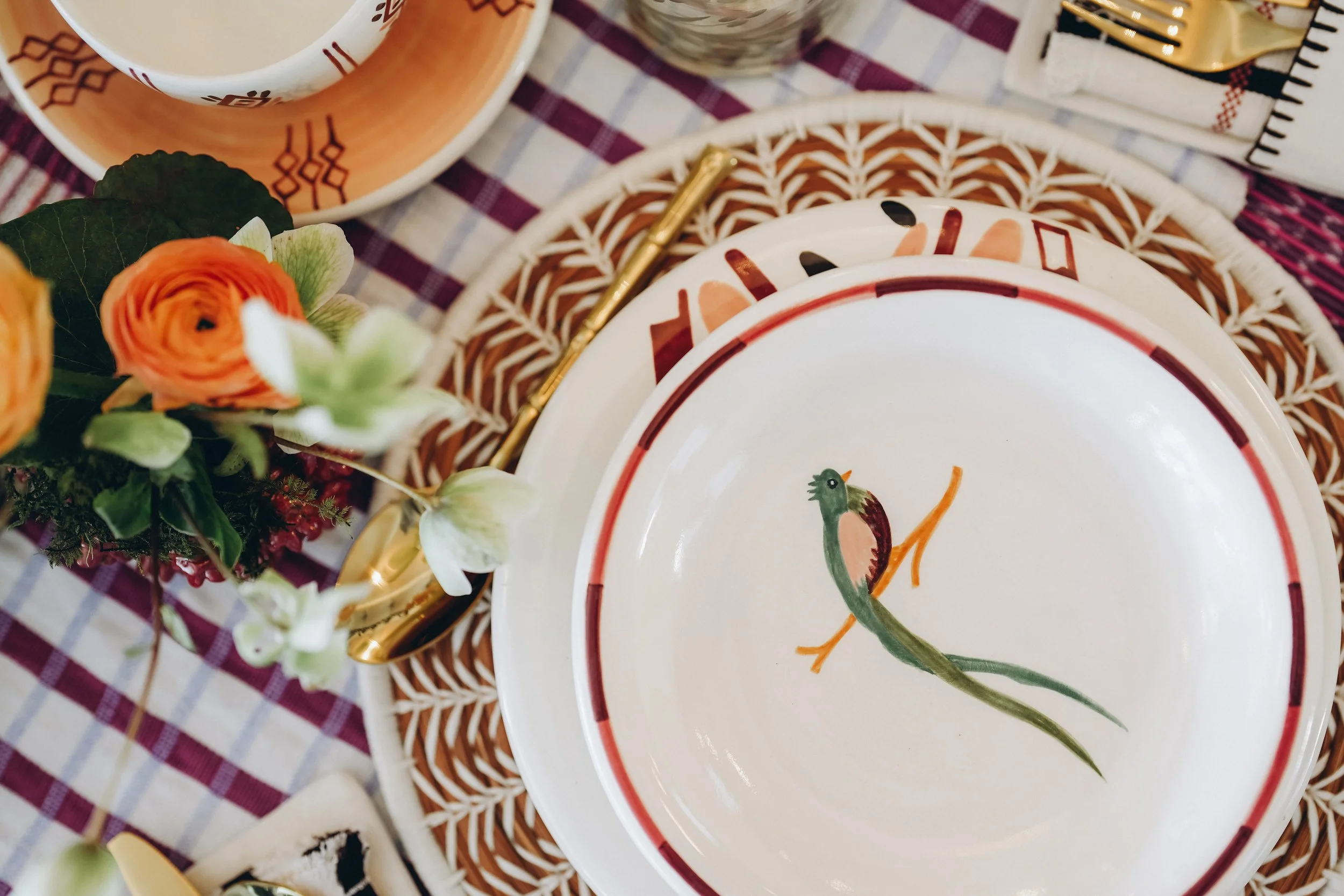 Hand-painted bird bowl layered with artisan ceramics, a pine needle woven charger, gold flatware, flowers, and RB Curated textiles.