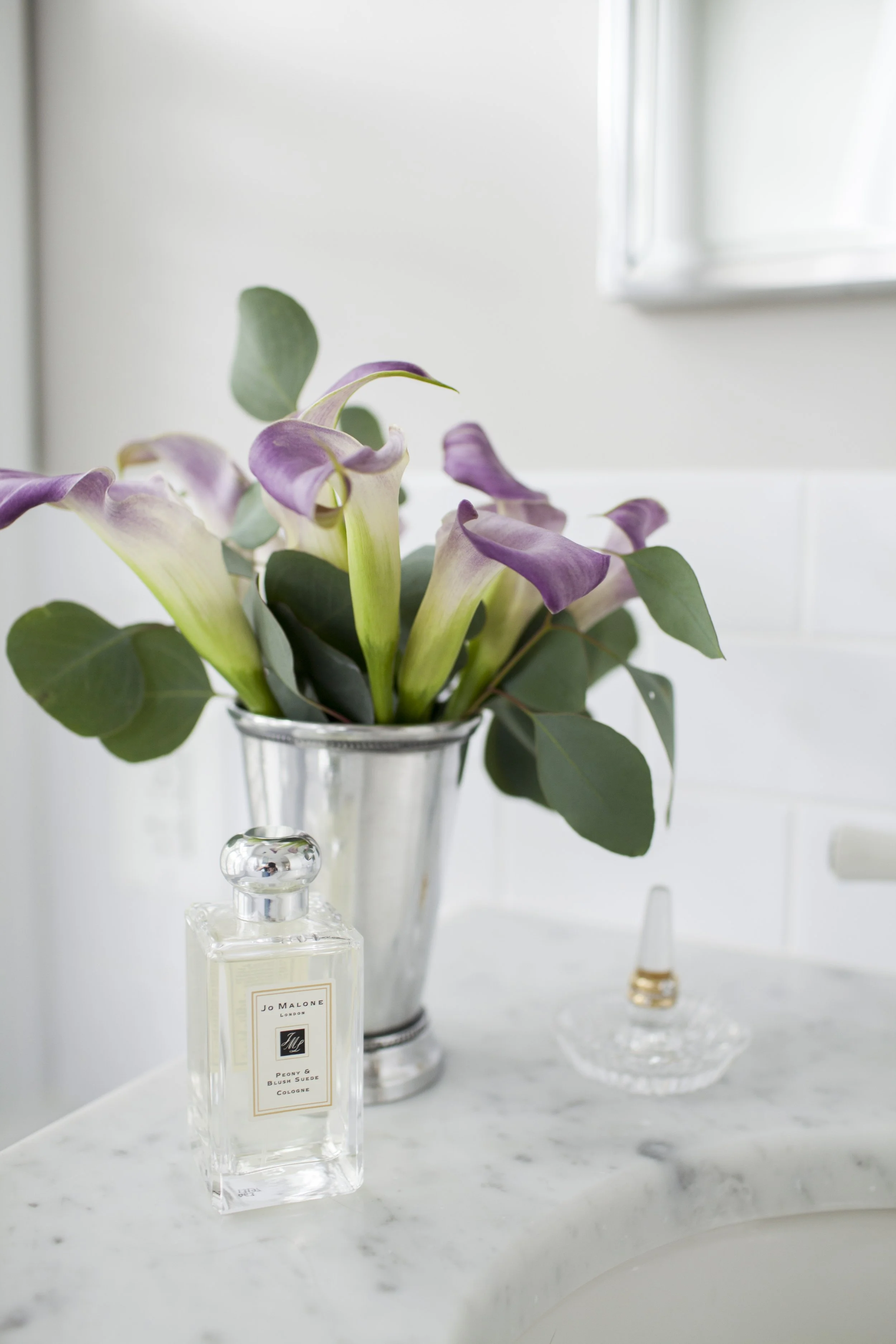 Purple calla lilies in a silver vase beside a perfume bottle on a marble bathroom countertop.