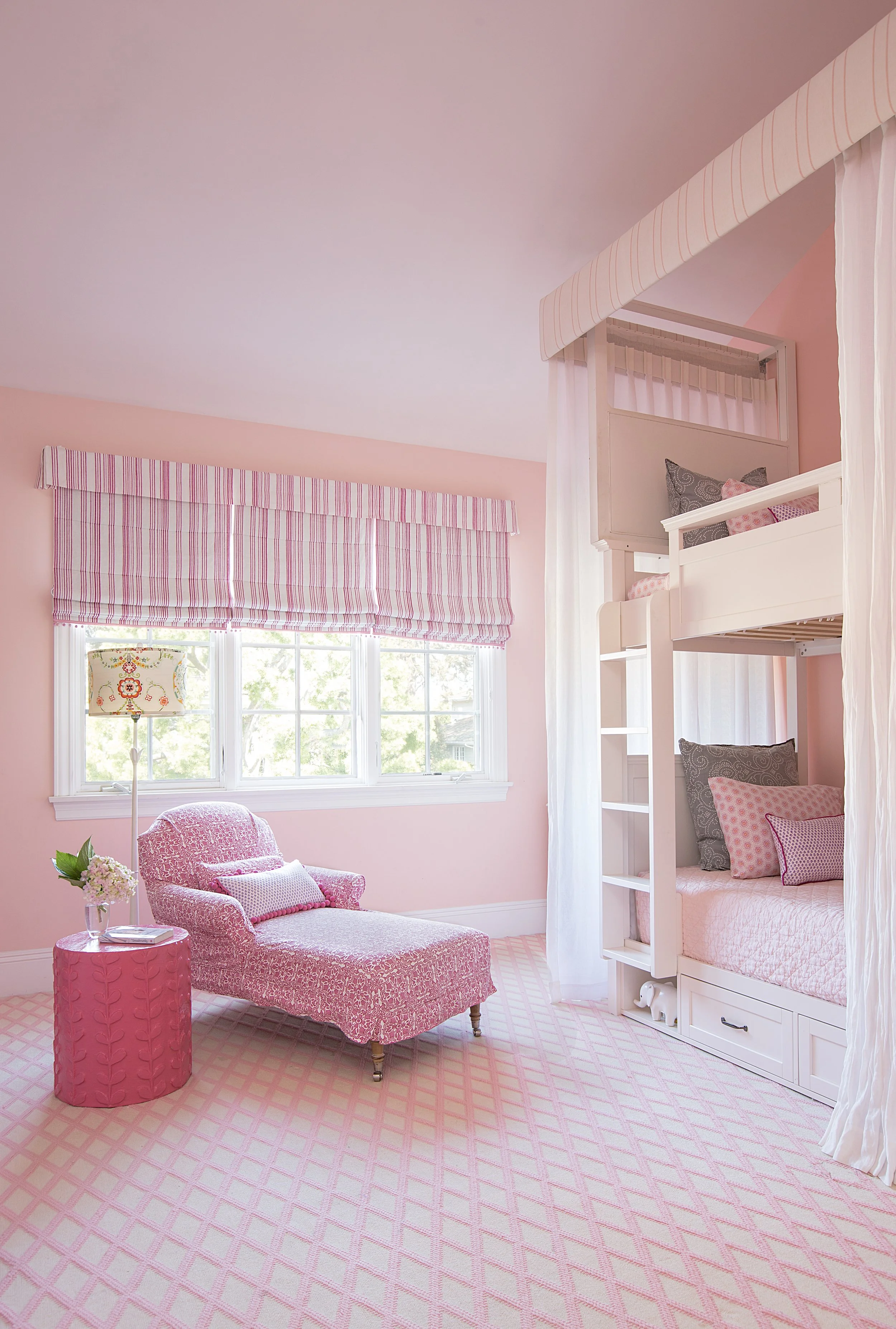 Playful pink bedroom with a patterned chaise, matching side table, floral lamp, bunk bed, and striped window shade.