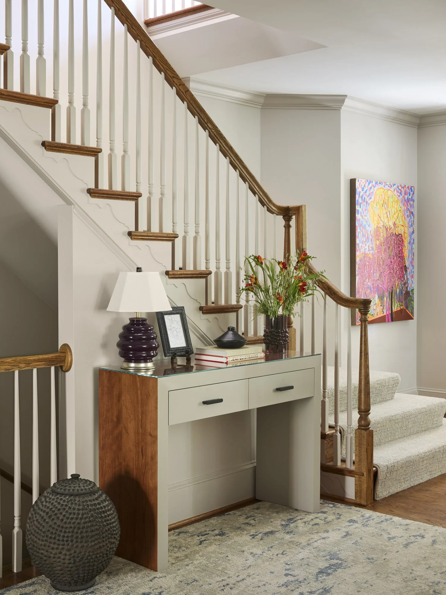 Entryway styled with a custom console, sculptural lamp, flowers, books, pottery, a patterned rug, and colorful artwork.