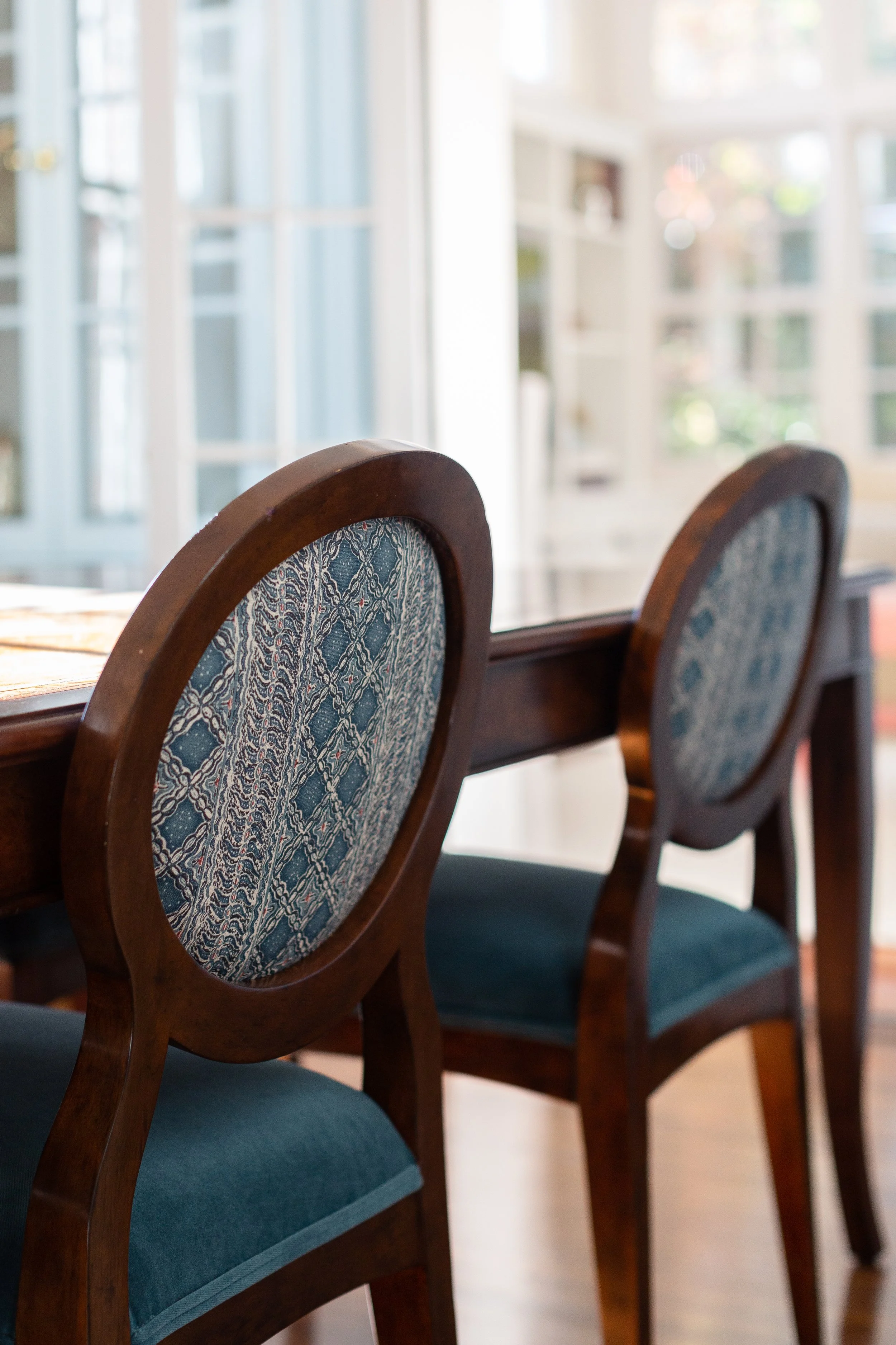 Close-up of blue upholstered wood-frame dining chairs beside a wood table in a bright room.