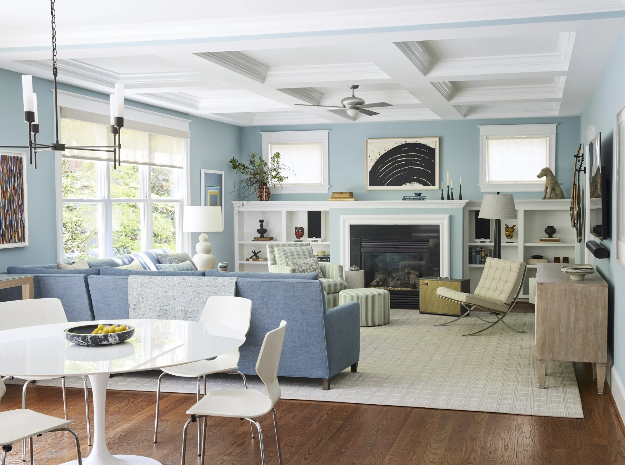 Light-filled living and dining room with pale blue cabinetry, a coffered ceiling, fireplace, and layered seating.