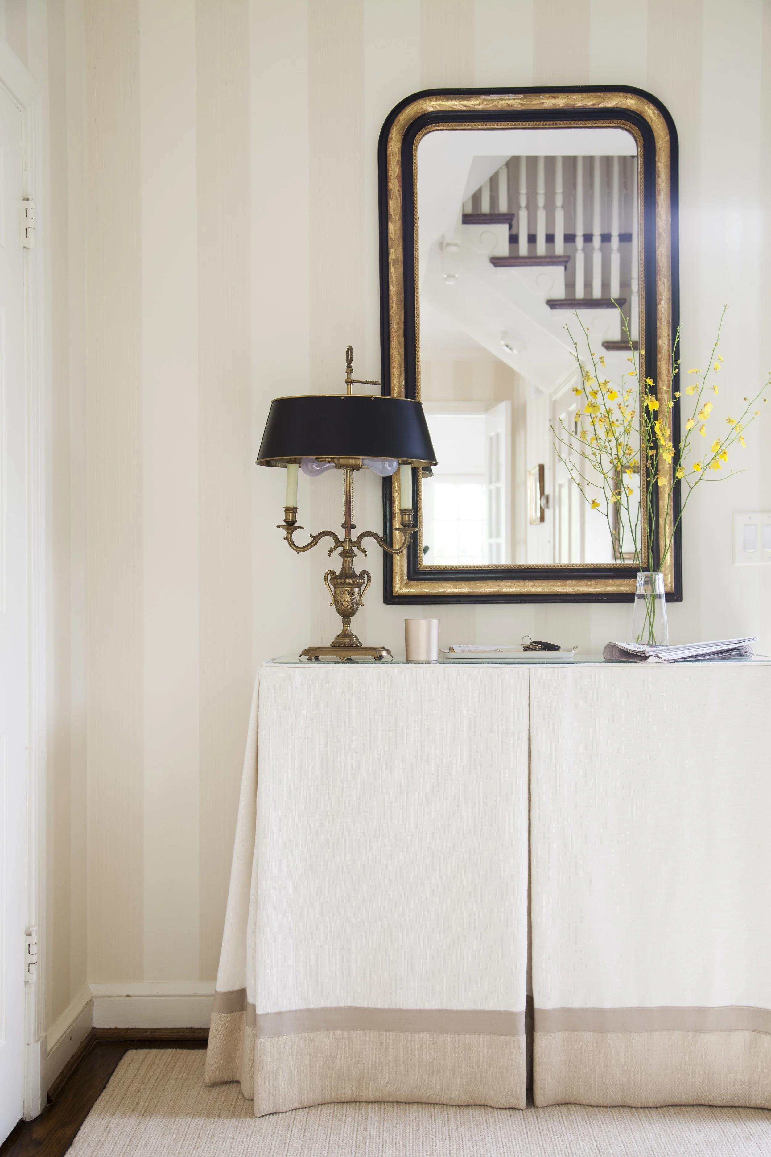 Friendship Heights entry hall with a large decorative mirror, skirted console table, vintage lamp, and vase of yellow flowers.