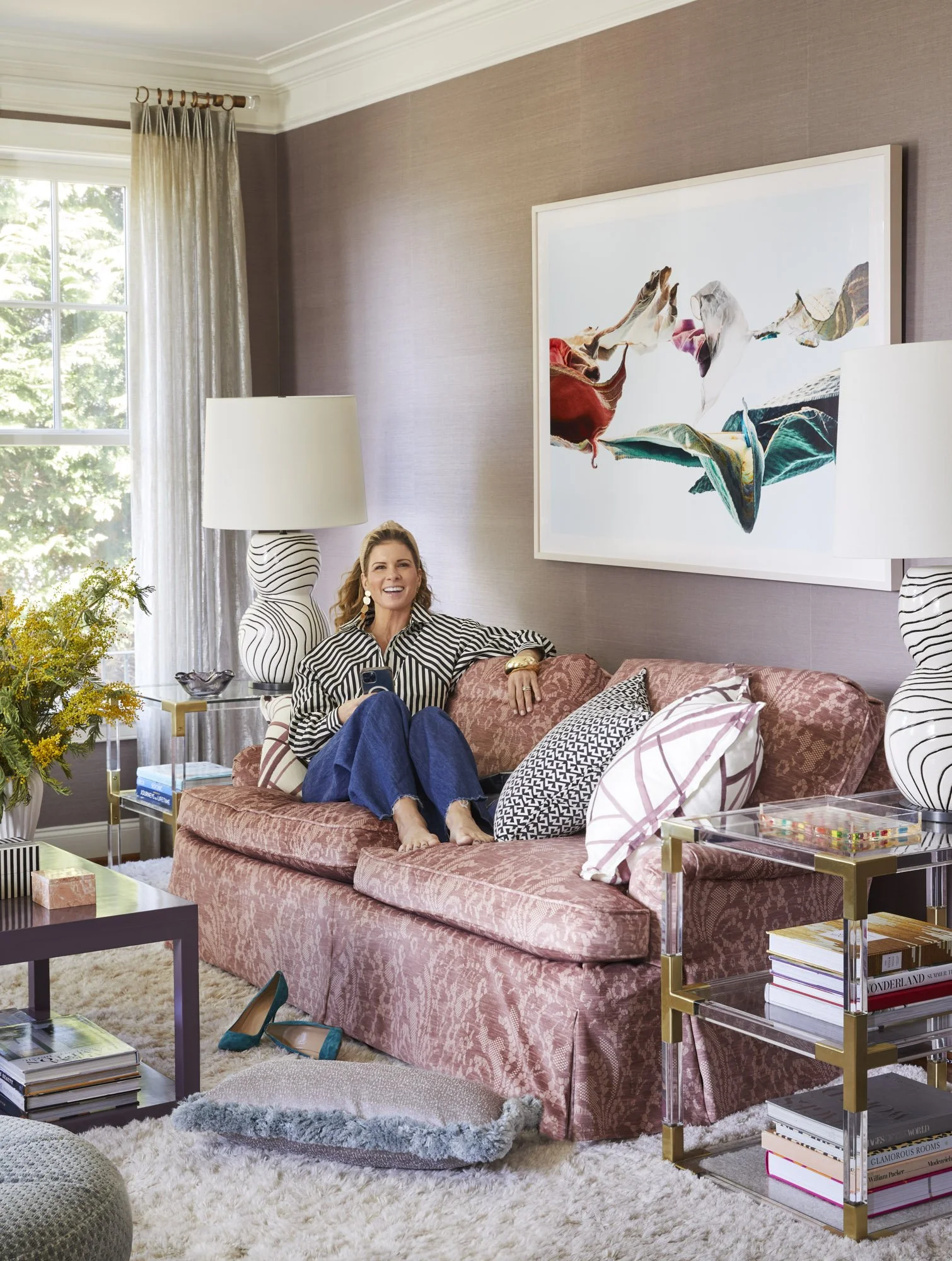 Woman seated on a pink patterned sofa beneath large abstract artwork in a living room with striped lamps and layered pillows.