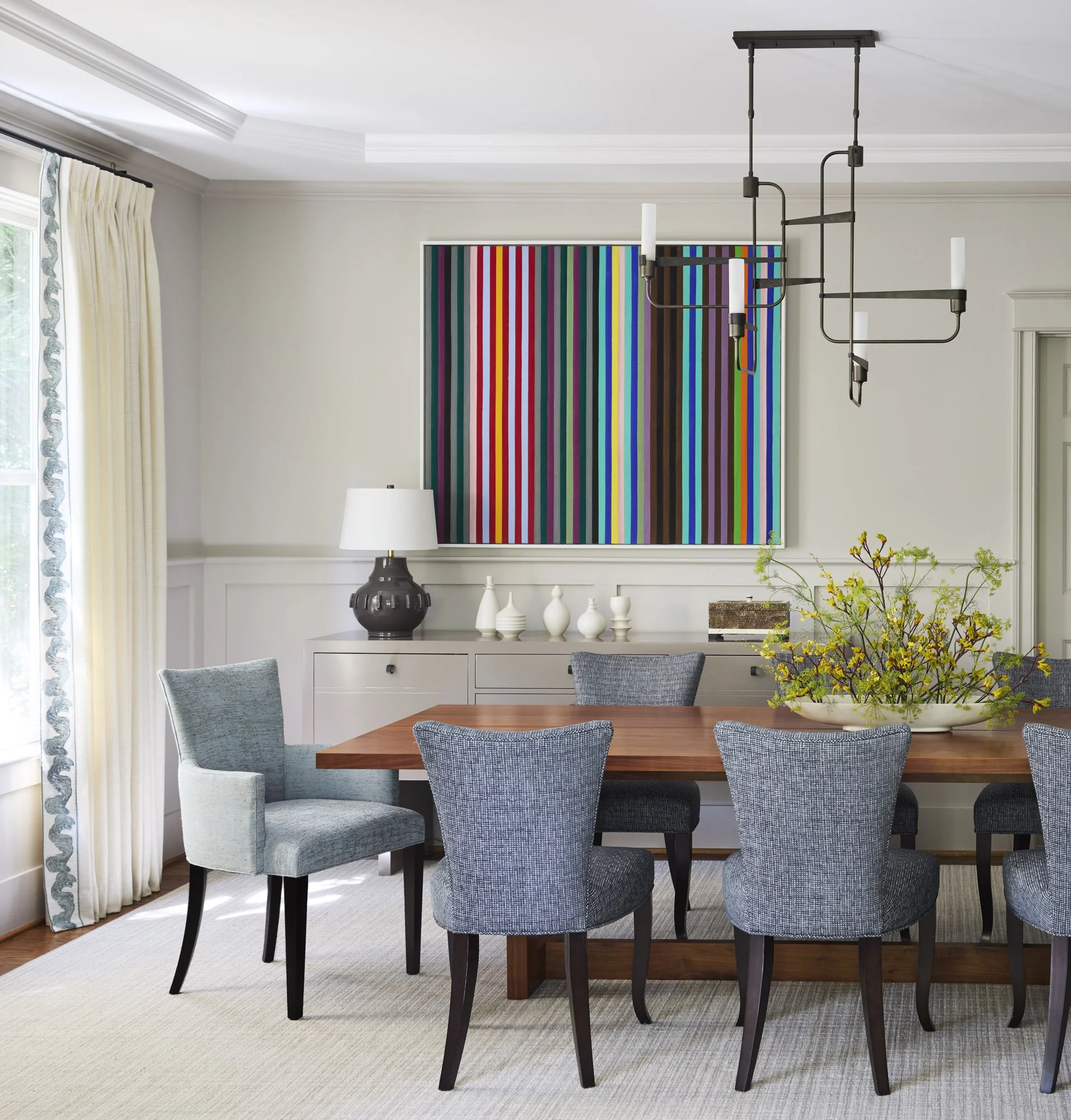Large abstract artwork displayed in a light-filled dining room with cane chairs and colorful flowers.