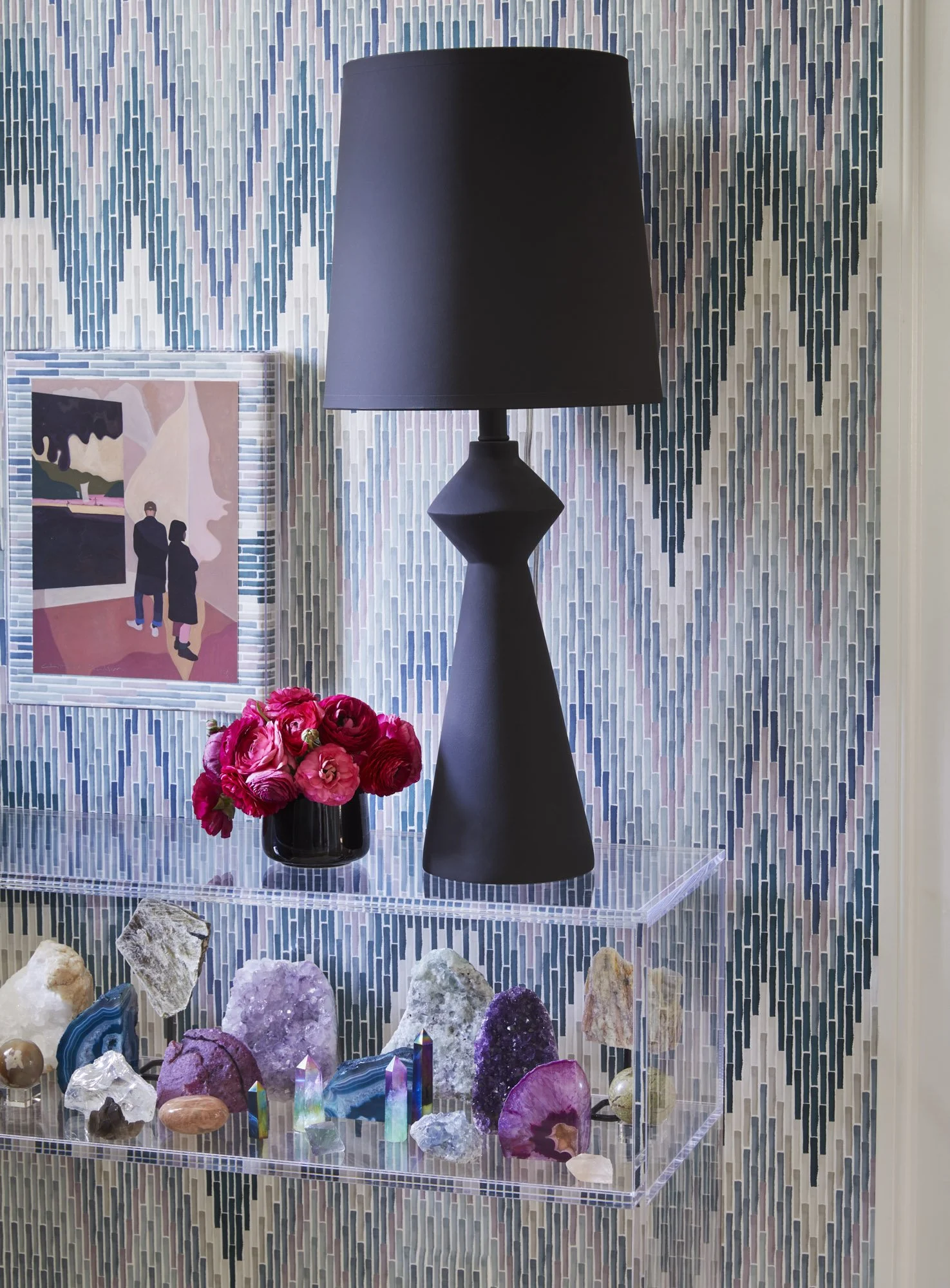 Styled shelf with a black lamp, small framed artwork, pink flowers, and colorful crystals against a patterned wall.