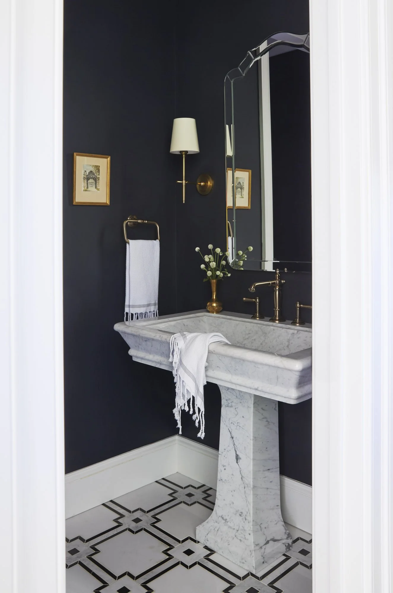 Dramatic powder room with dark walls, a marble pedestal sink, brass fixtures, framed artwork, and patterned floor tile.
