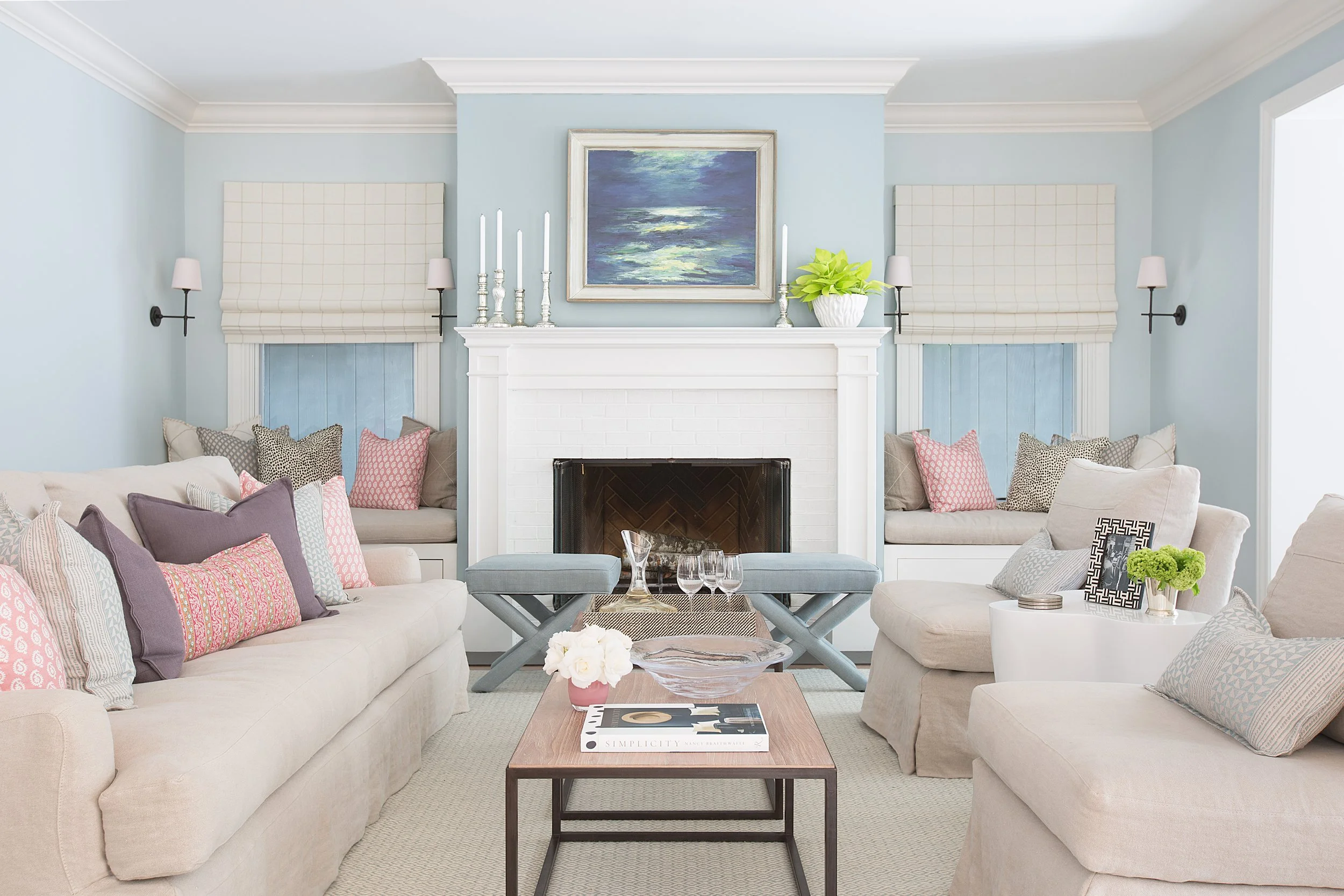 Light-filled living room with pale blue walls, a white fireplace, beige sofas, patterned pillows, and a central coffee table.