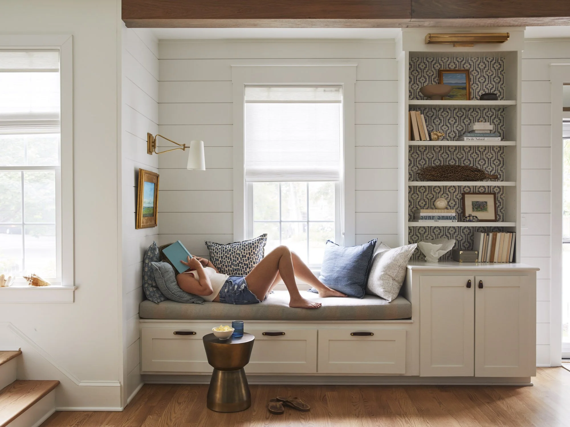 Person reads in a built-in window nook surrounded by patterned pillows, bookshelves, and natural light.