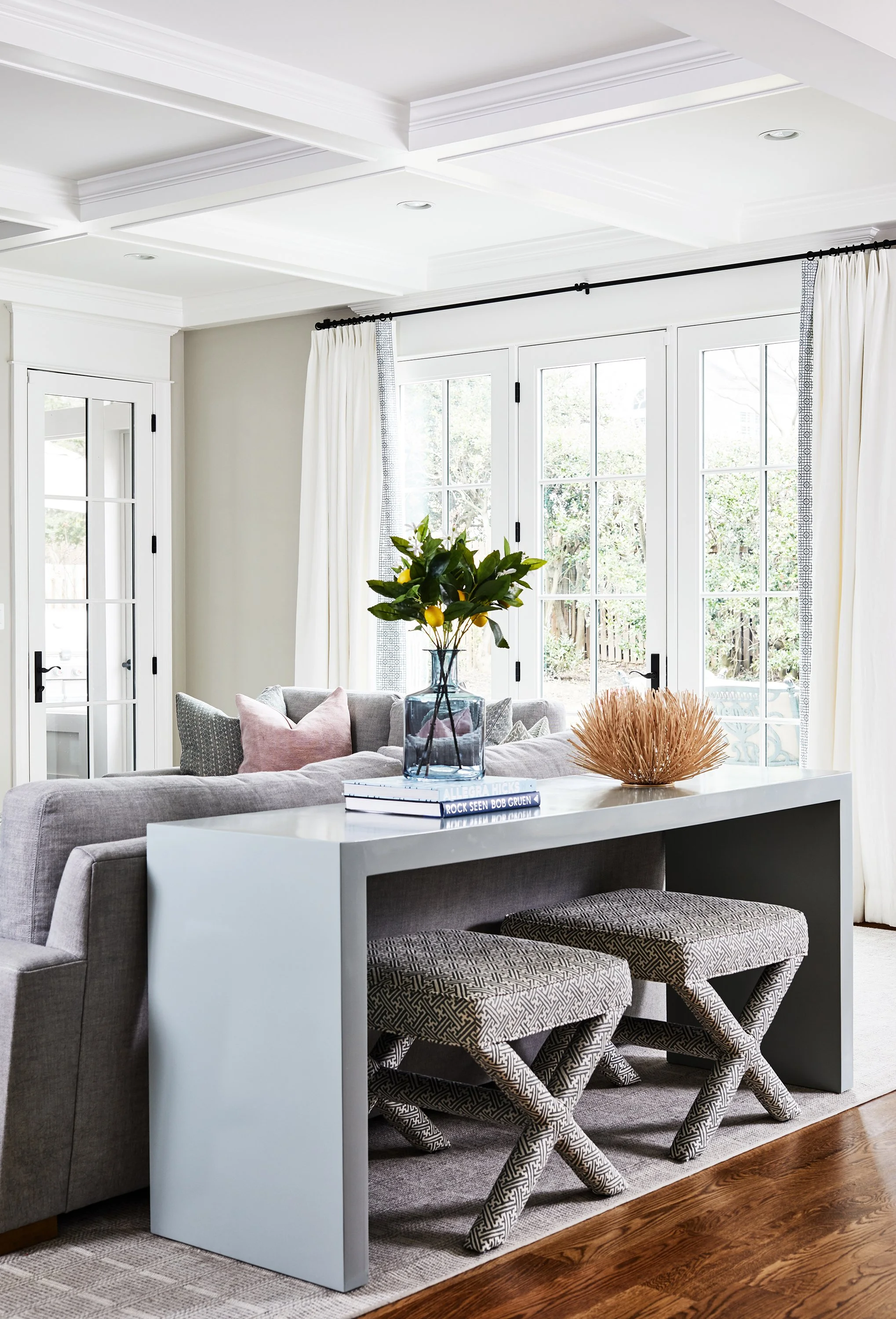 Gray sofa backed by a slim console table with decorative objects and two patterned stools near large windows.