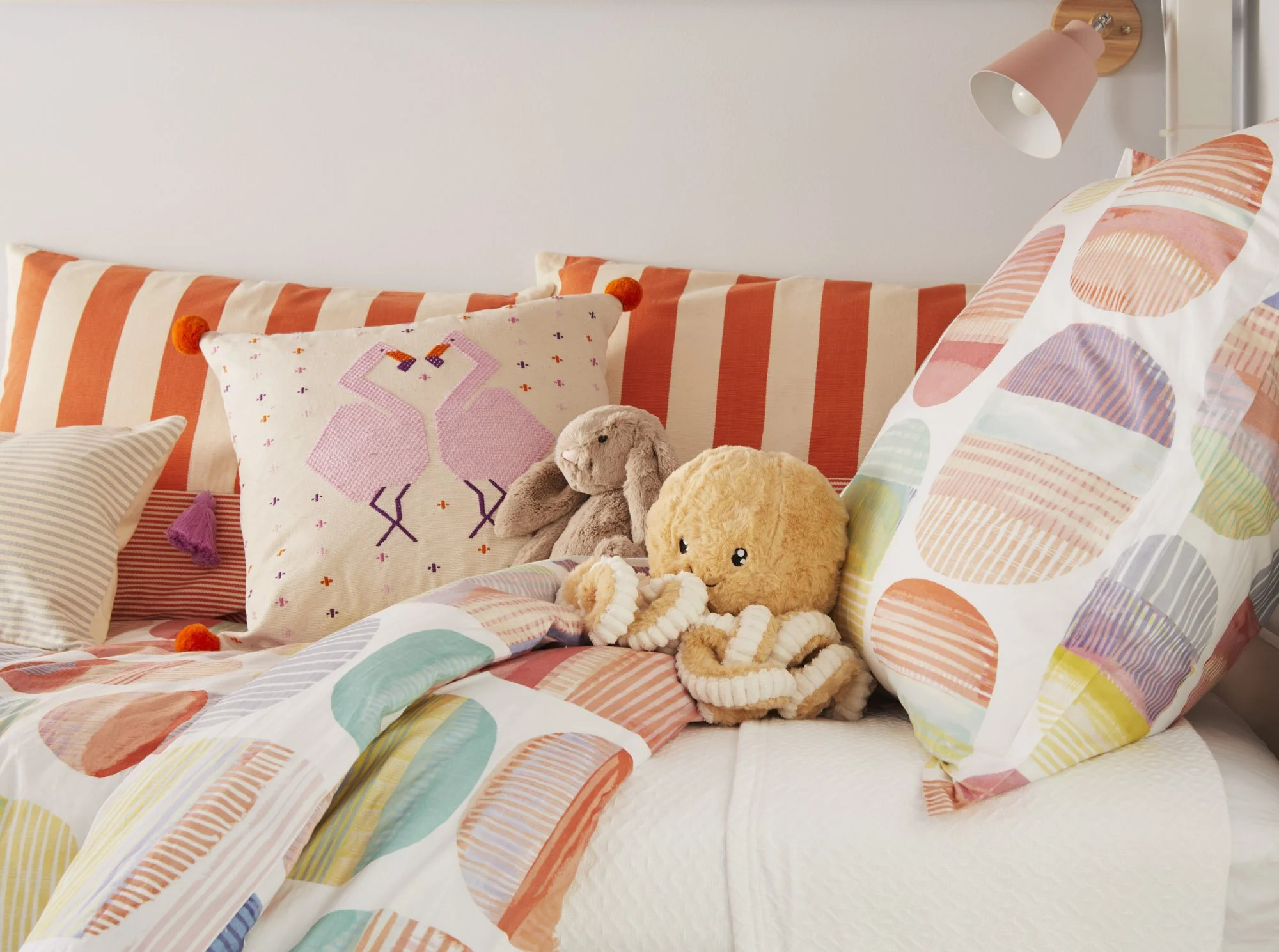 Playful girls’ bedroom with colorful patterned bedding, an RB Curated flamingo pillow, stuffed animals, and a wall-mounted light.