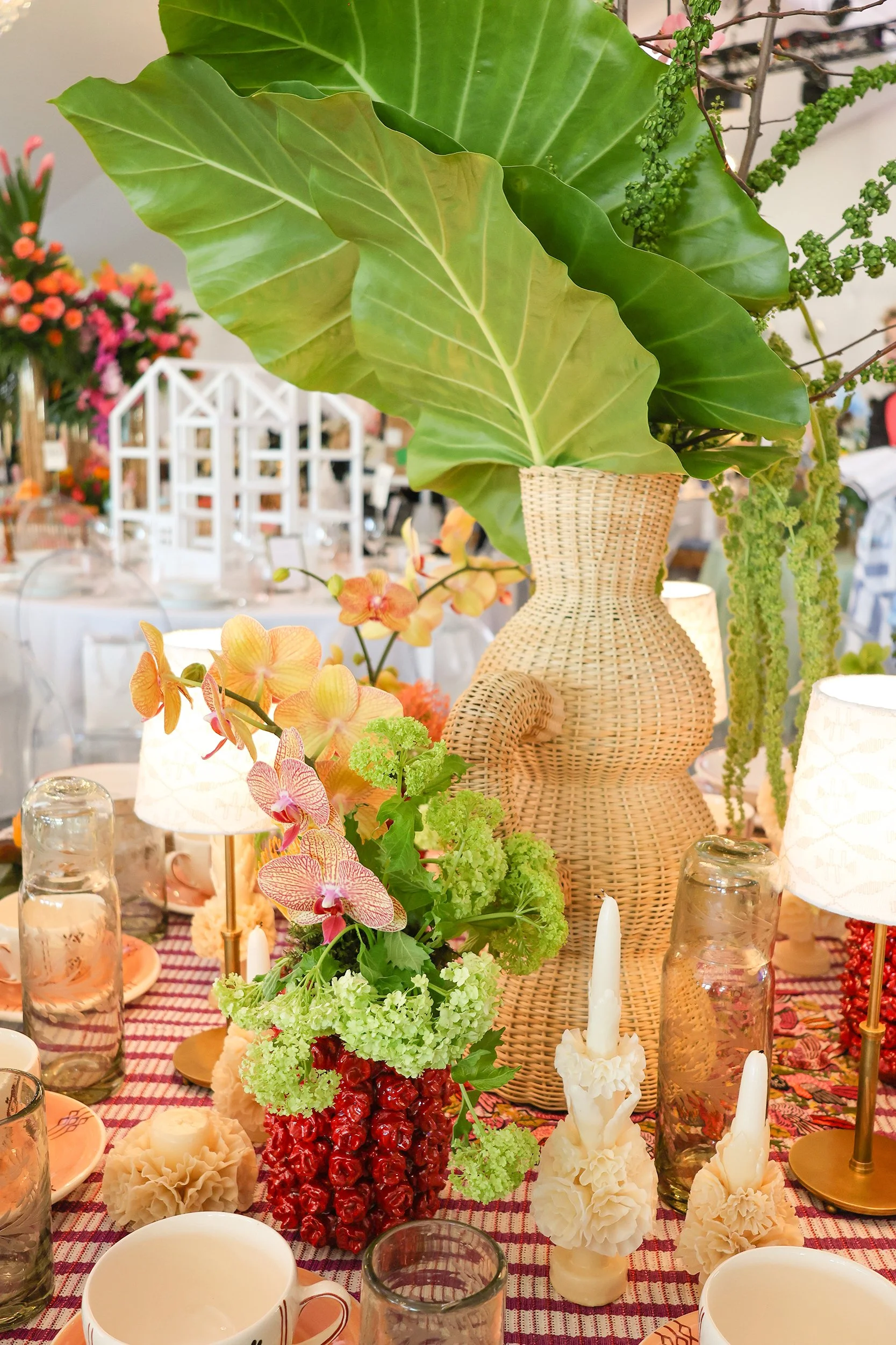 Naples Tables centerpiece with oversized tropical leaves, a woven vase, orchids, colorful flowers, candles, and handmade tableware.
