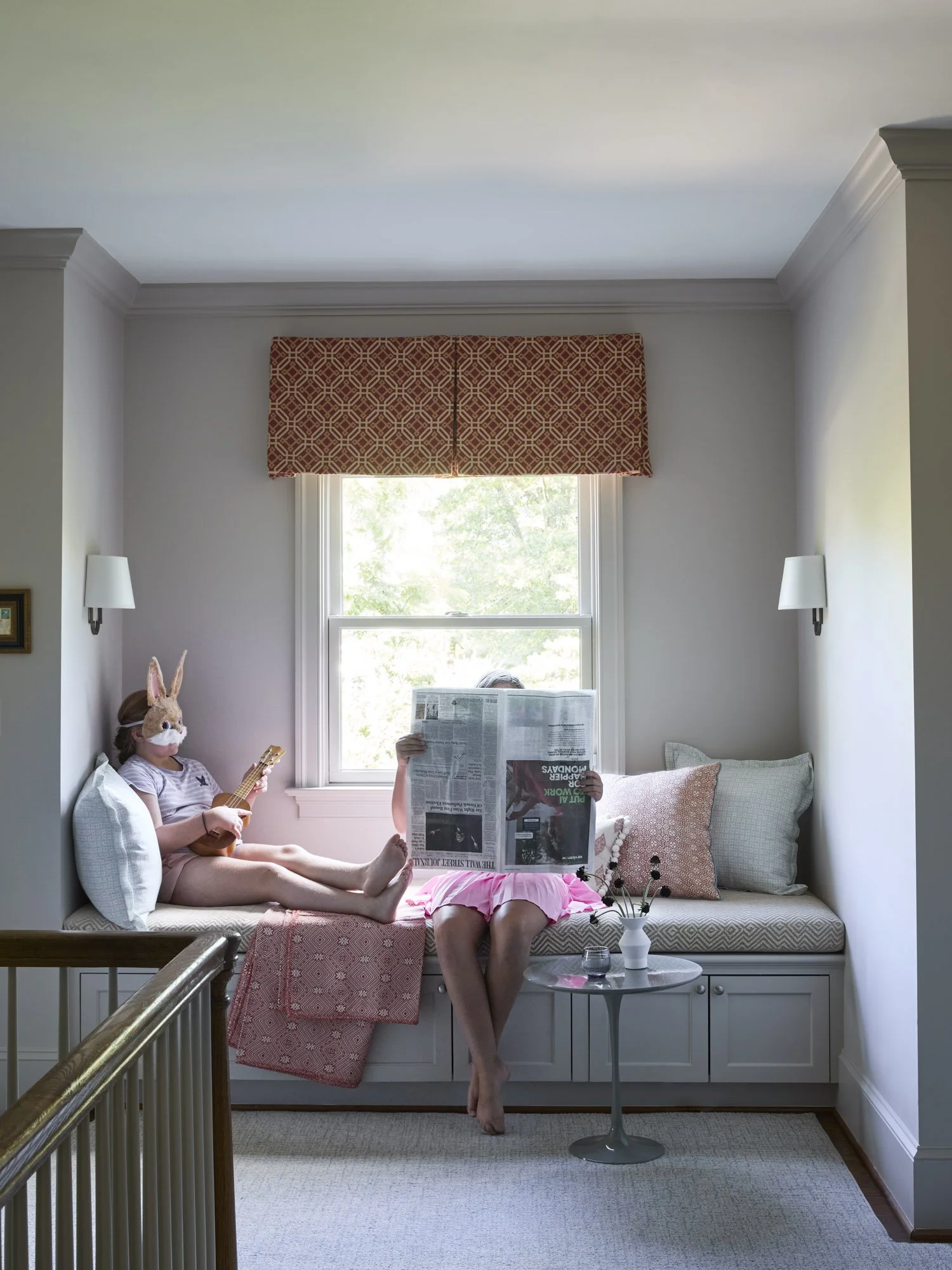 Two people relax on a patterned window seat, one reading and the other wearing a rabbit mask while holding a ukulele.