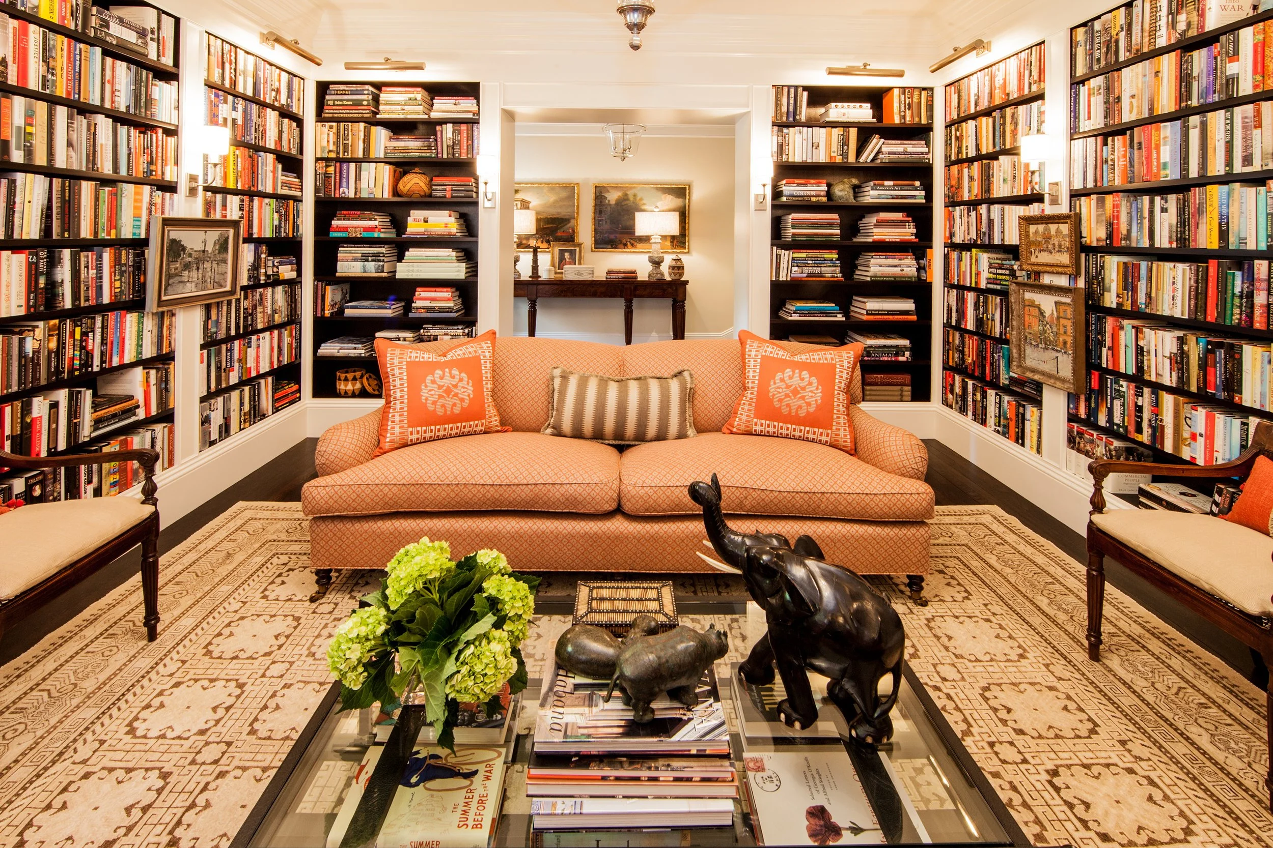 Home library with an orange sofa, floor-to-ceiling bookshelves, glass coffee table, elephant sculpture, and green hydrangeas.