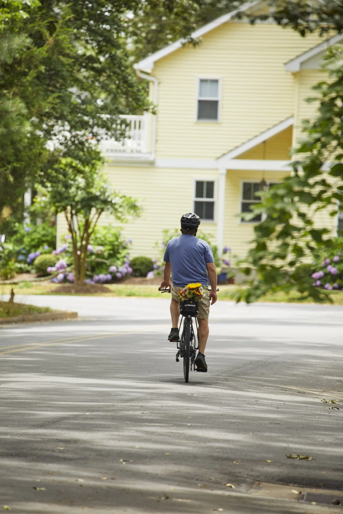 Person rides a bicycle along a tree-lined Rehoboth Beach street bordered by hydrangeas and a yellow home.