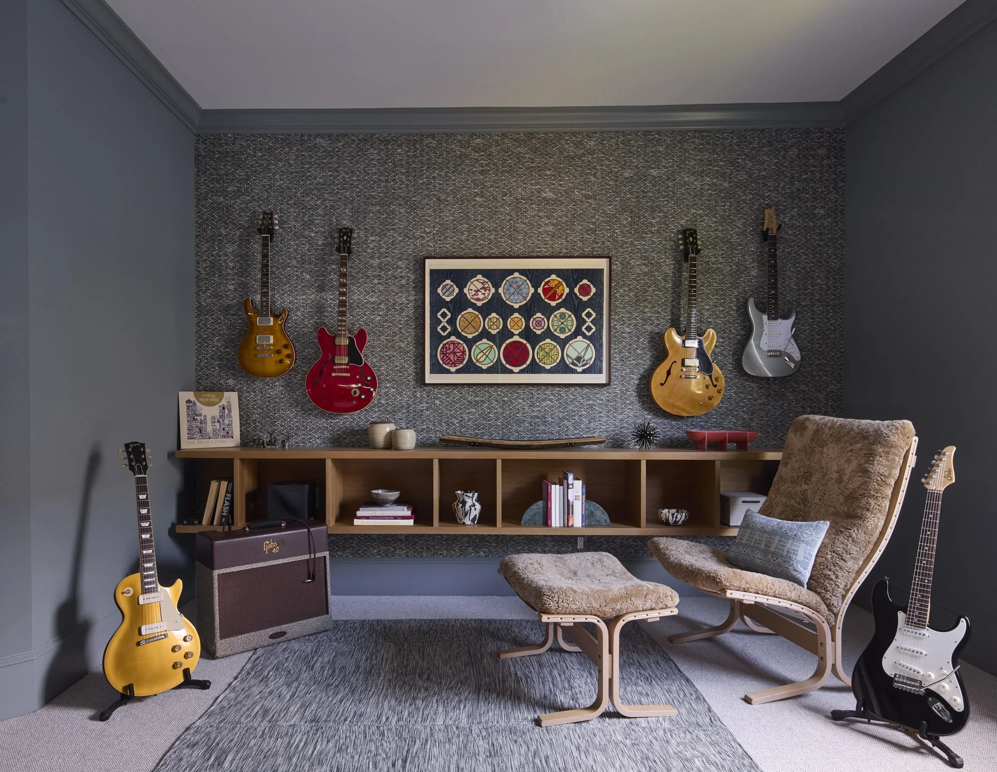 Music room with wall-mounted guitars, a comfortable chair and ottoman, amplifier, books, collected objects, and a patterned rug.