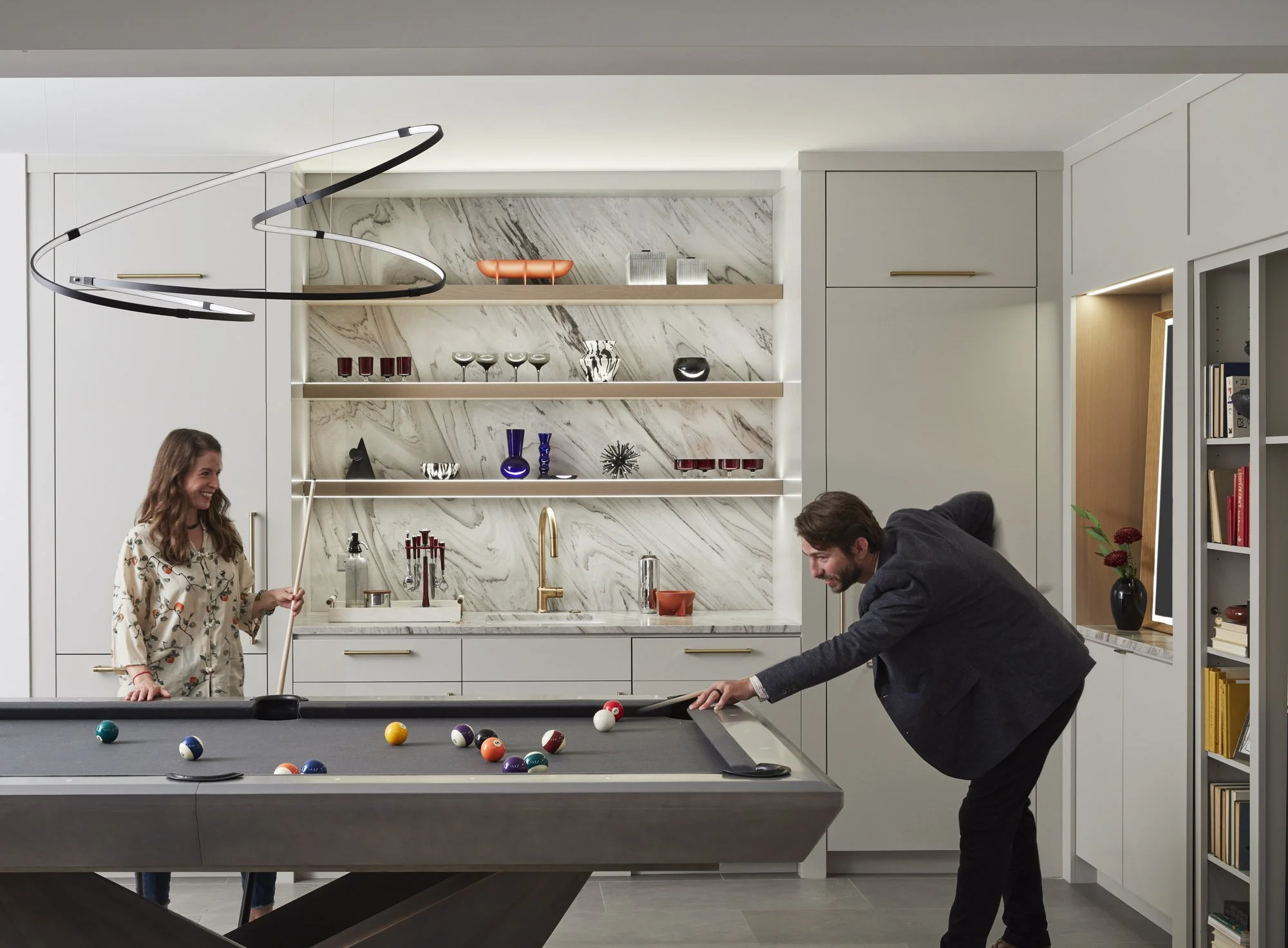 Family members play pool in a modern billiards room with custom cabinetry, open shelving, and a marble backsplash.