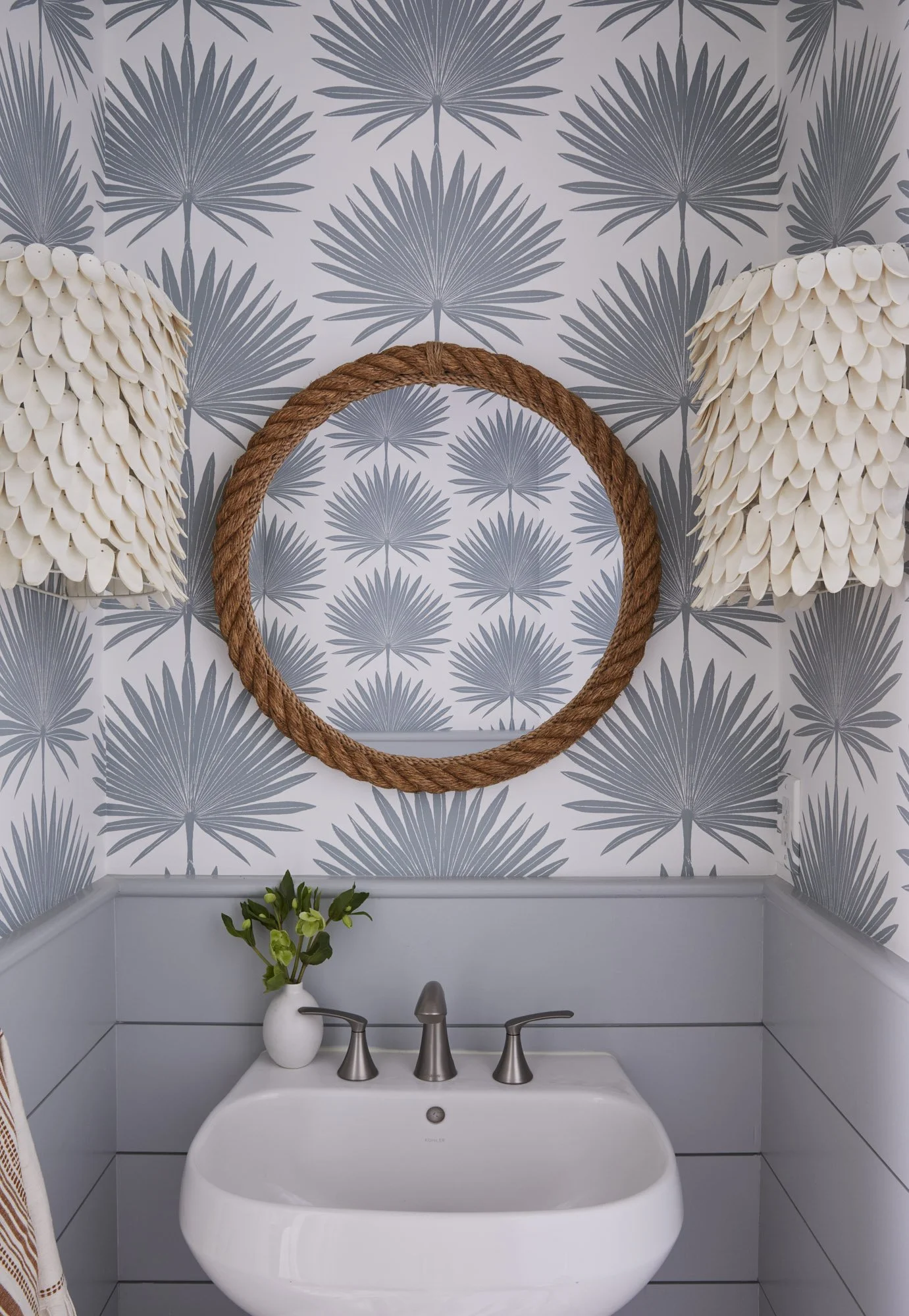 Coastal bathroom with palm-leaf wallpaper, a rope-framed mirror, shell-patterned pendant lights, and a white sink.
