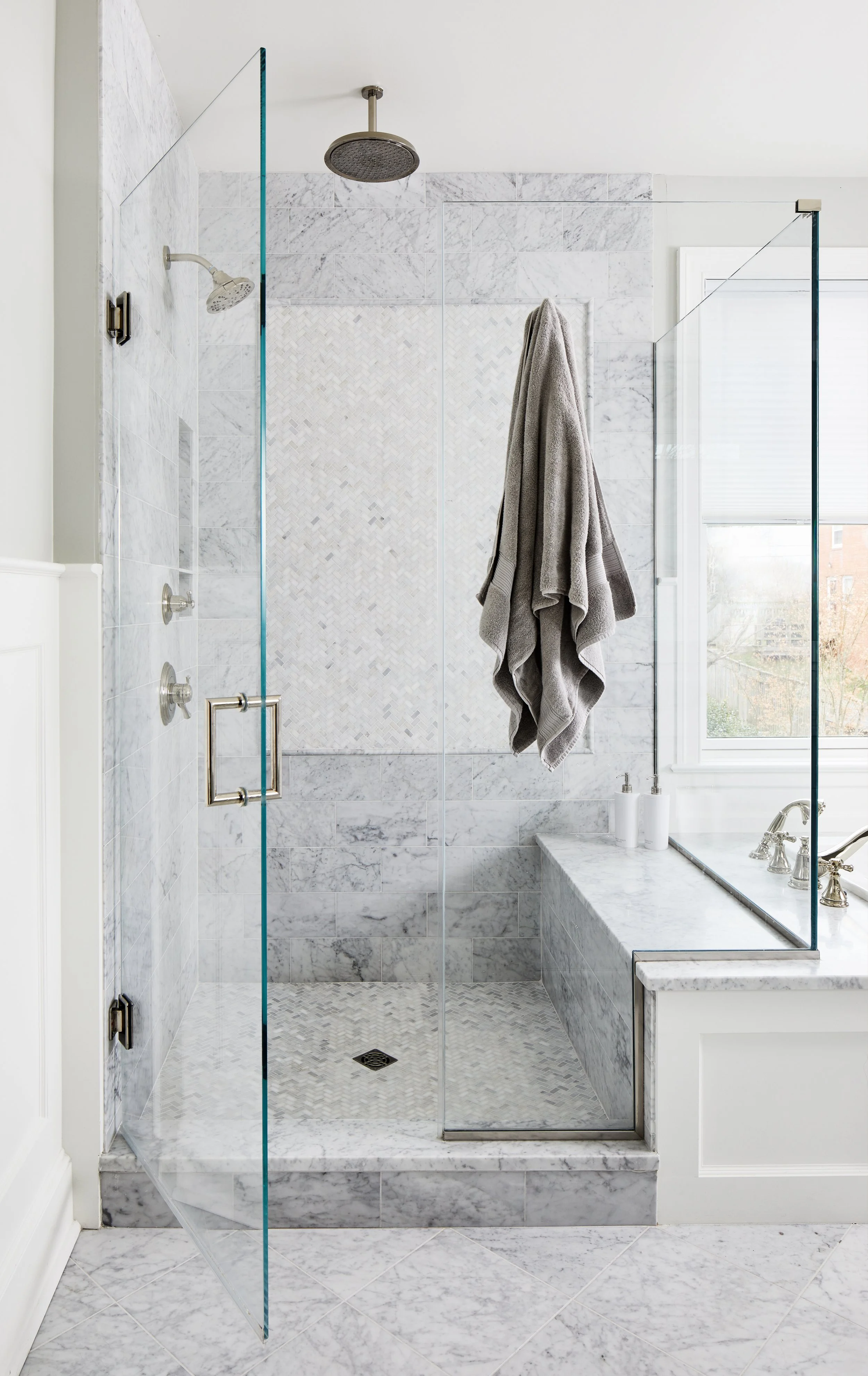 White marble shower with a glass enclosure, silver fixtures, rainfall showerhead, and patterned floor tile.