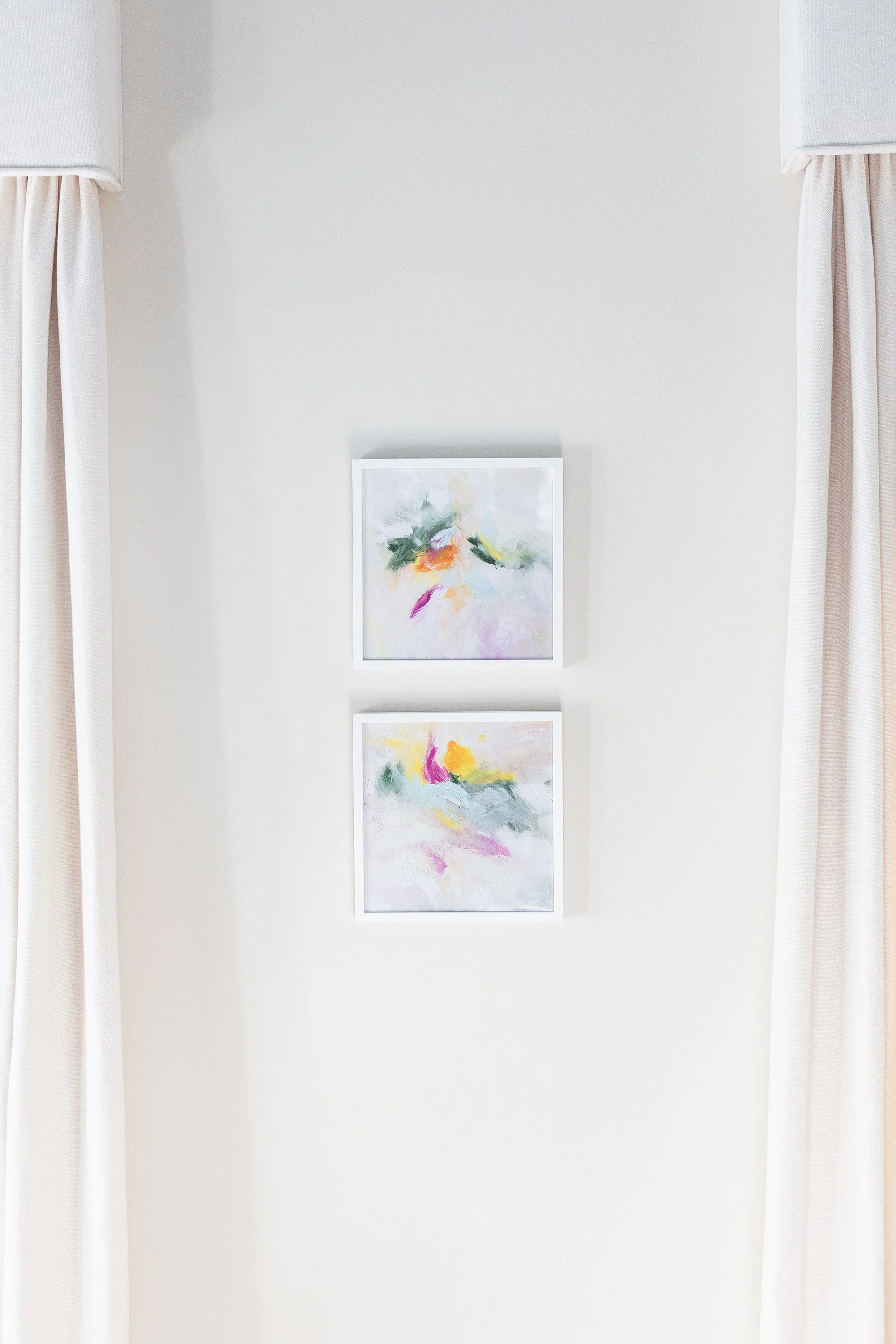 Two sunlit windows framed by white curtains with pale blue trim, with small abstract paintings displayed between them.