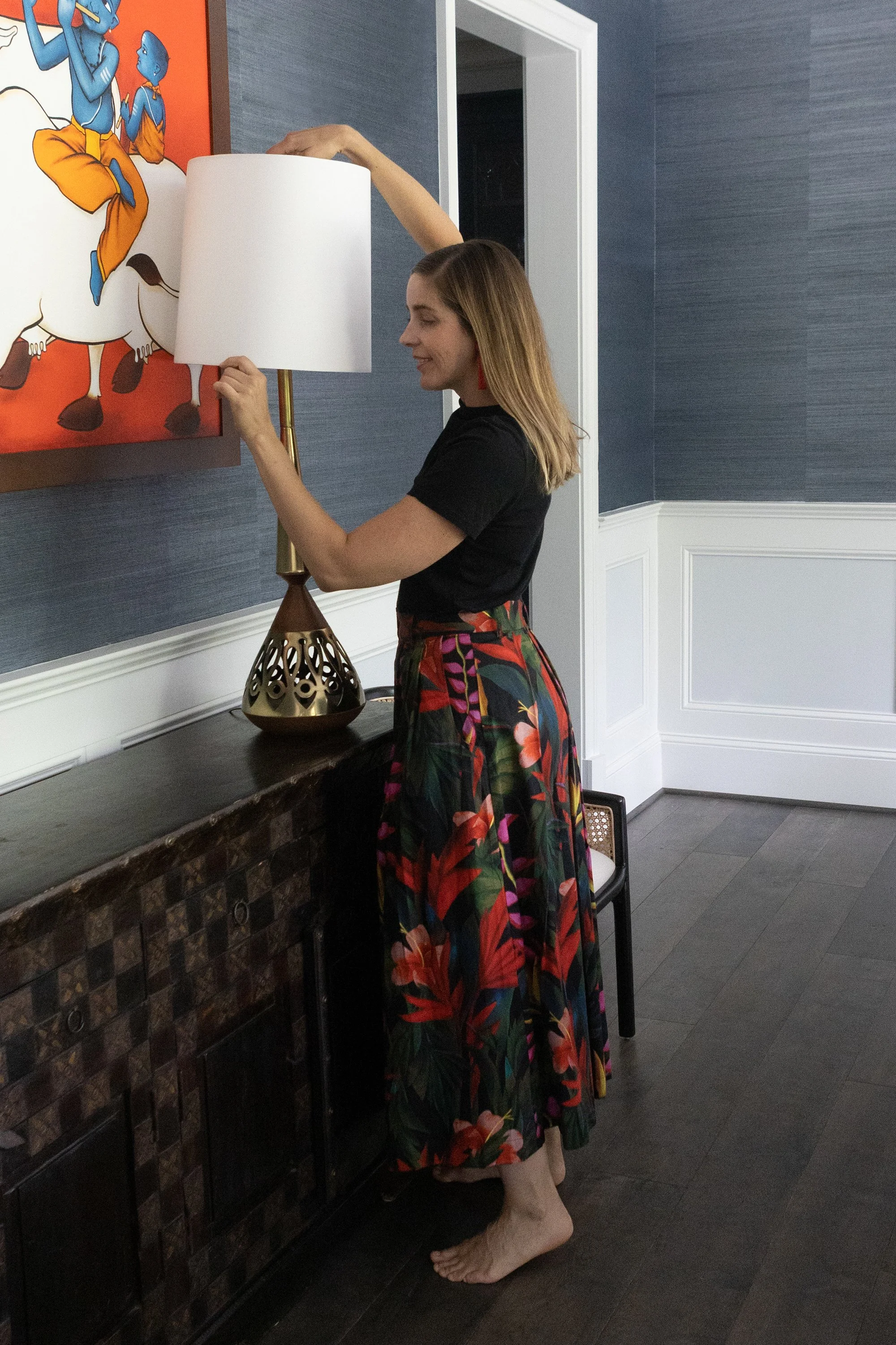 Regan adjusting a table lamp on a dining room sideboard beneath colorful abstract artwork.
