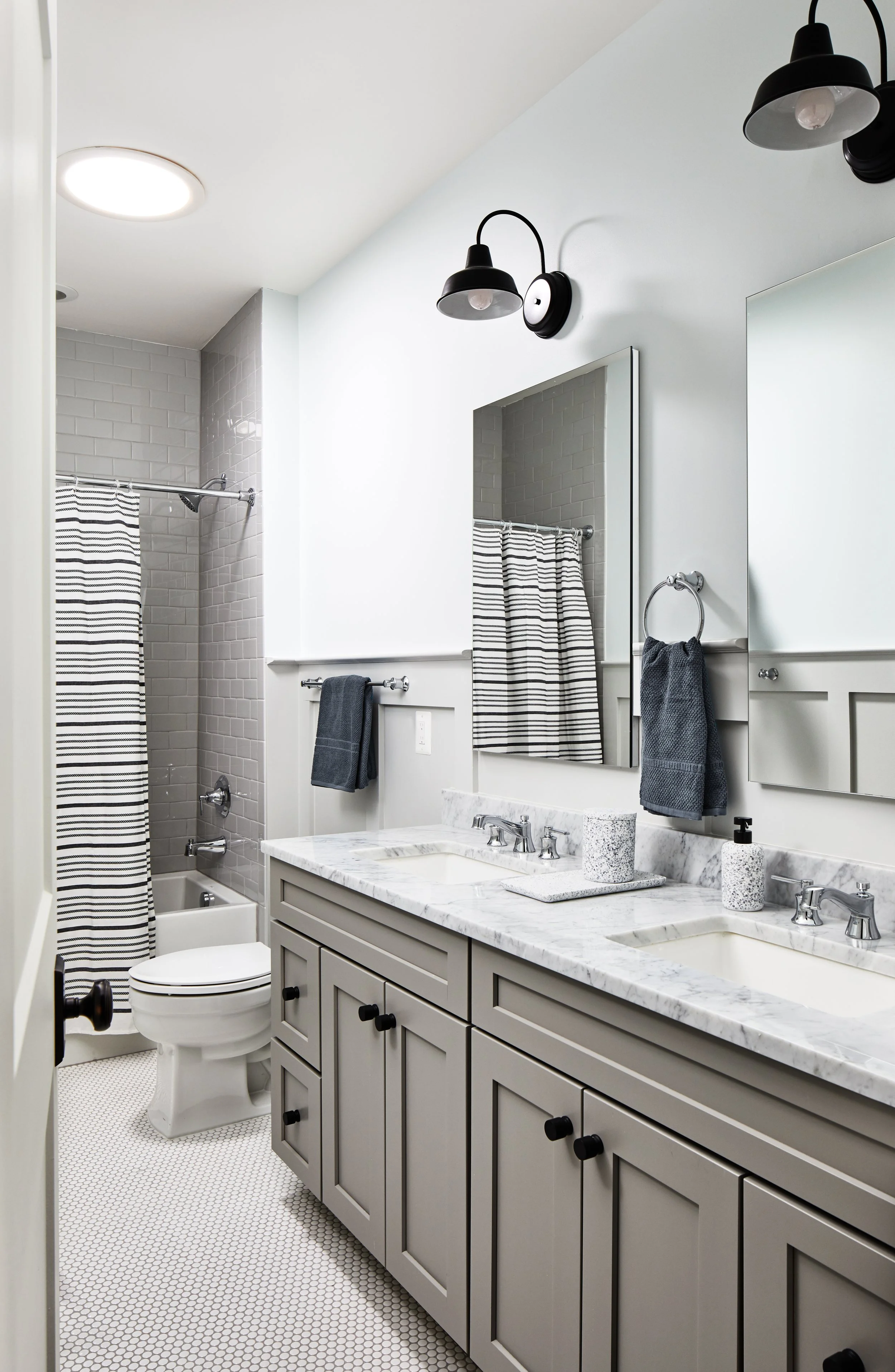 Modern bathroom with dual sinks, light gray cabinets, marble countertop, and tile flooring. Features a toilet, bathtub with a striped shower curtain, and wall-mounted lighting fixtures.
