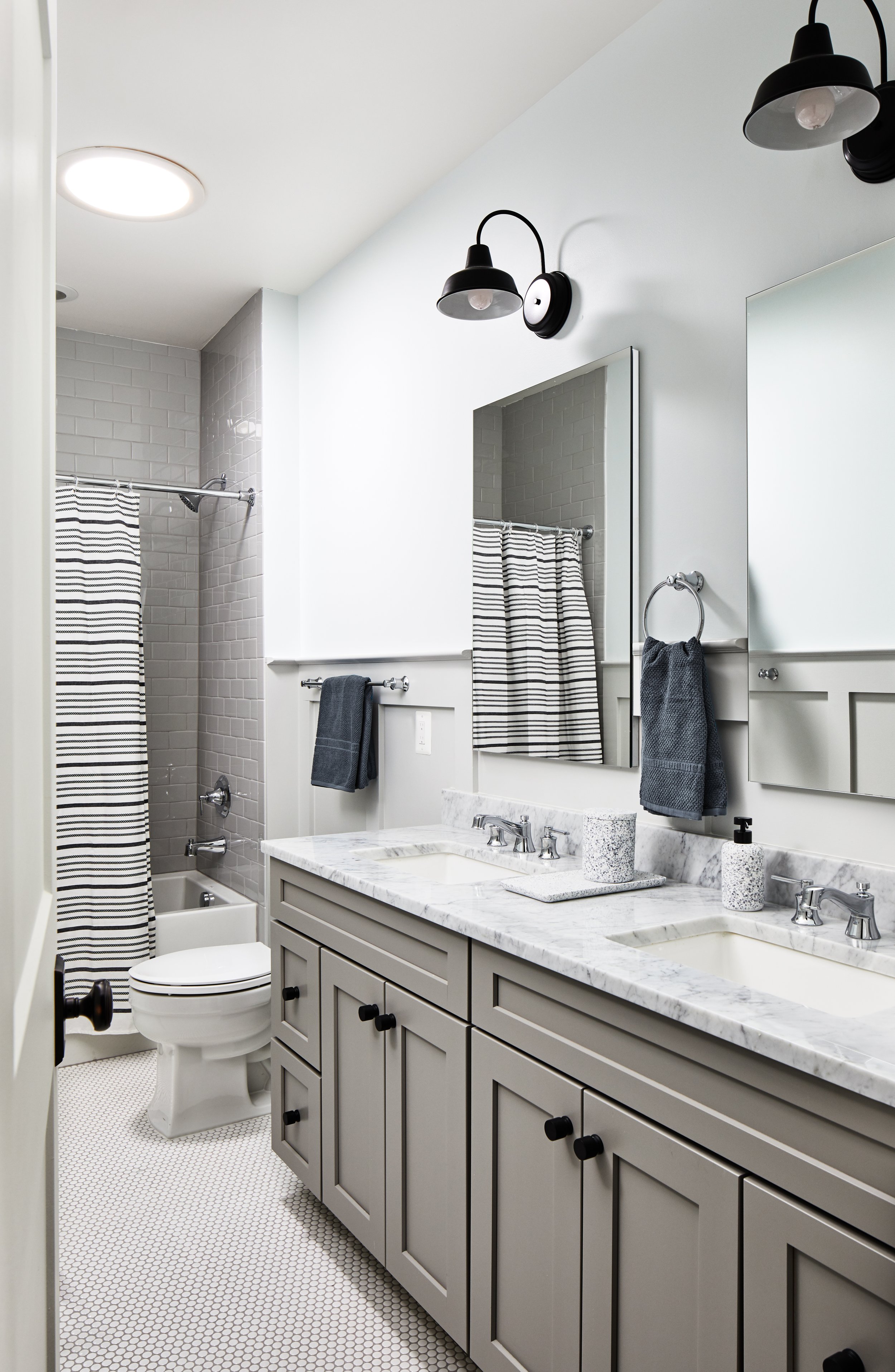 Family bathroom with two sinks, light gray cabinetry, marble countertop, bathtub, striped shower curtain, and wall-mounted lights.