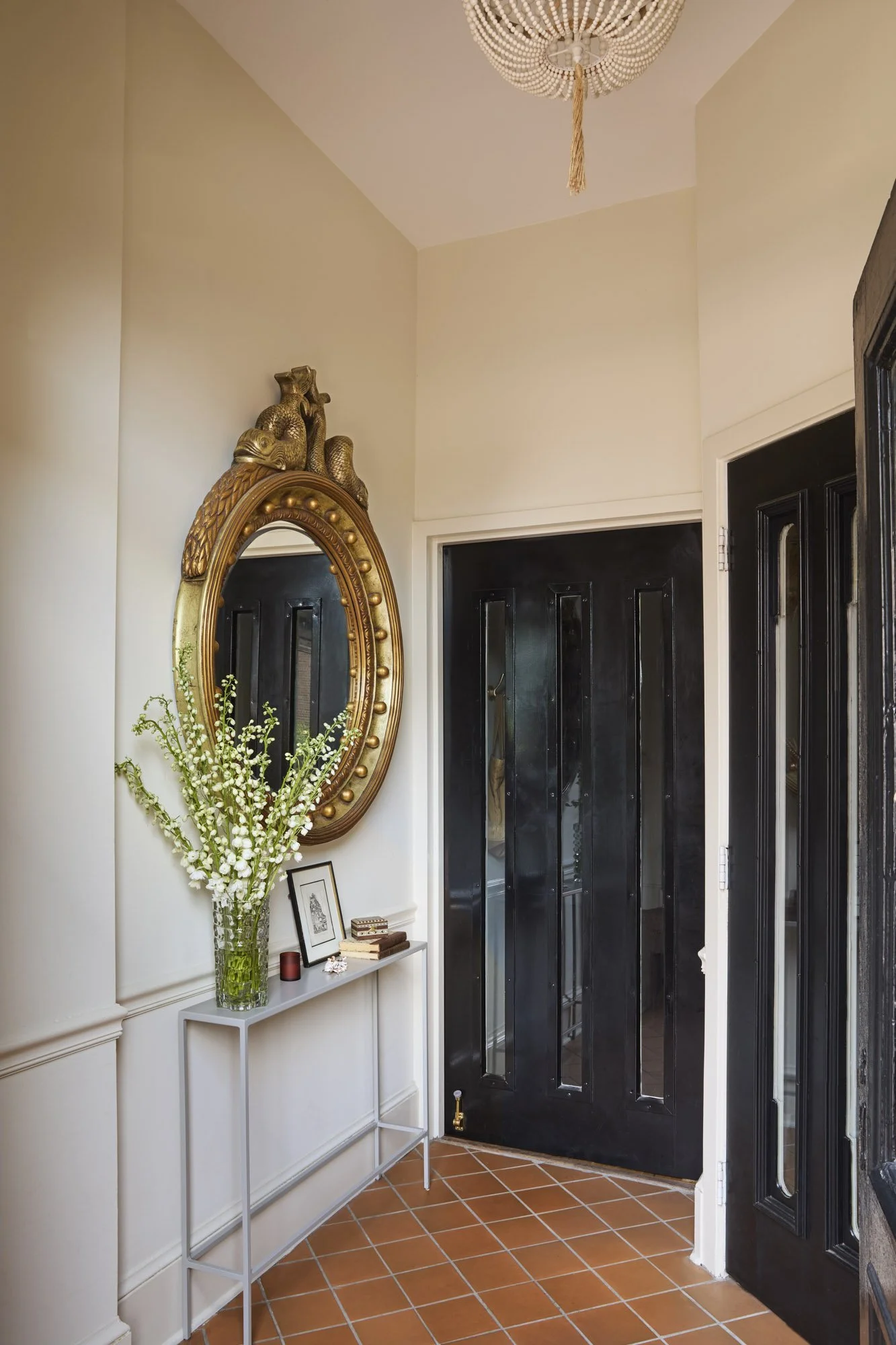 RBI Studio entryway with black doors, a sculptural gold mirror, white flowers, terracotta flooring, and a beaded chandelier.