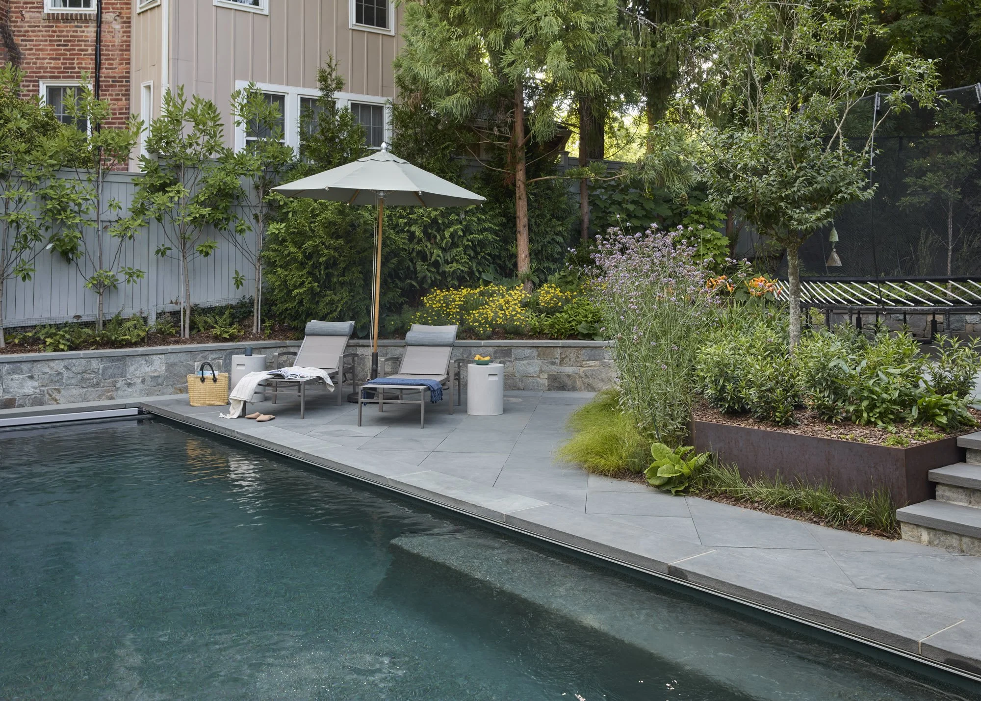 Landscaped backyard pool with a stone patio, lounge chairs, flowering gardens, and a raised planting bed.