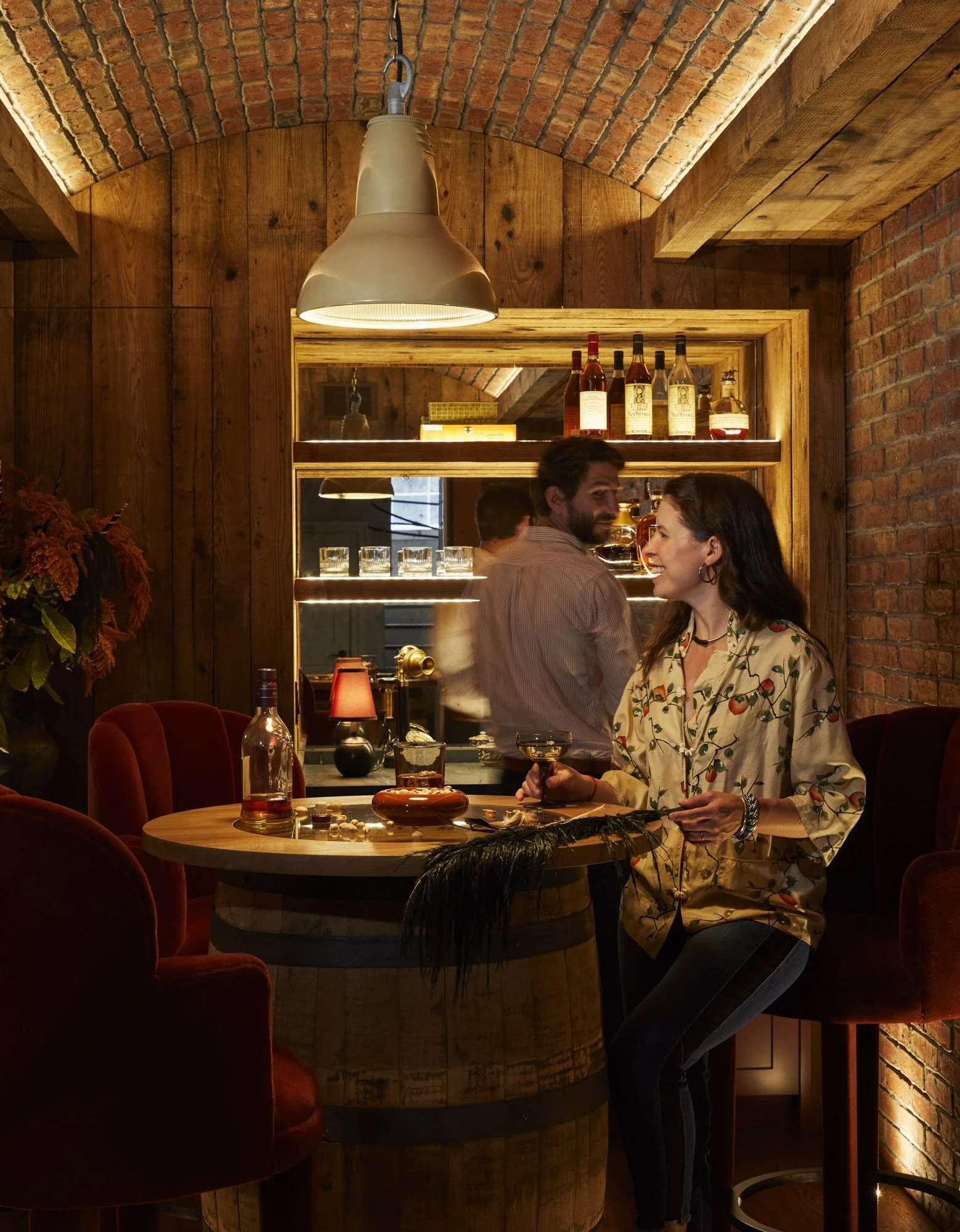 Couple enjoying drinks in the hidden speakeasy with brick walls, a barrel table, red seating, and illuminated bottle shelves.
