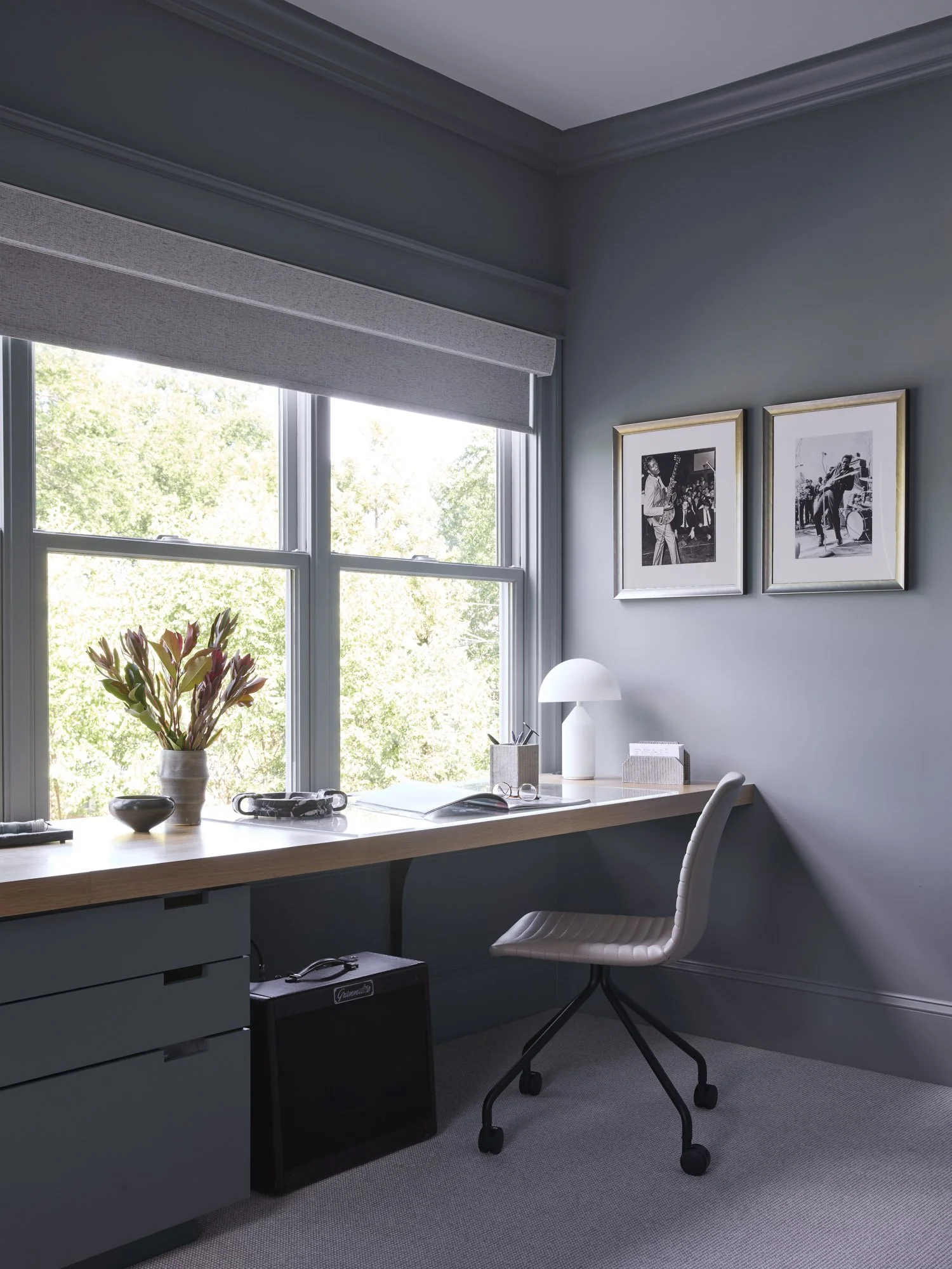 Neutral home office with a wood desk, rolling chair, large window, plants, framed photographs, and a decorative lamp.