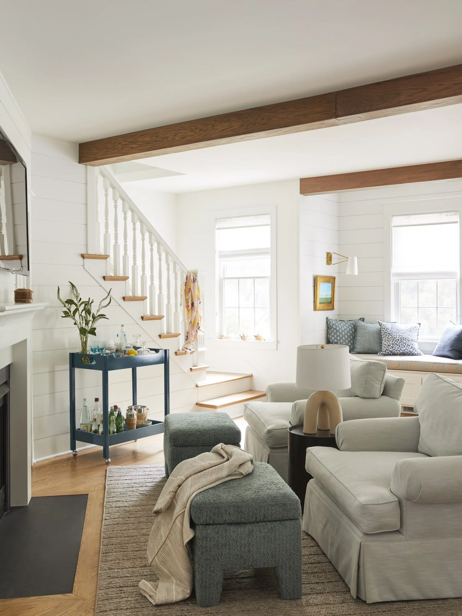 Light-filled beach house living room with a white sofa, fireplace, blue bar cart, exposed beams, staircase, and window seat.