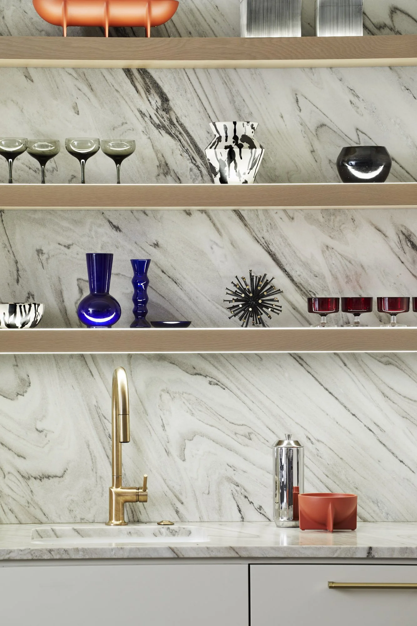 Custom bar with a marble backsplash, gold faucet, and open shelves displaying glassware, artwork, and collected objects.