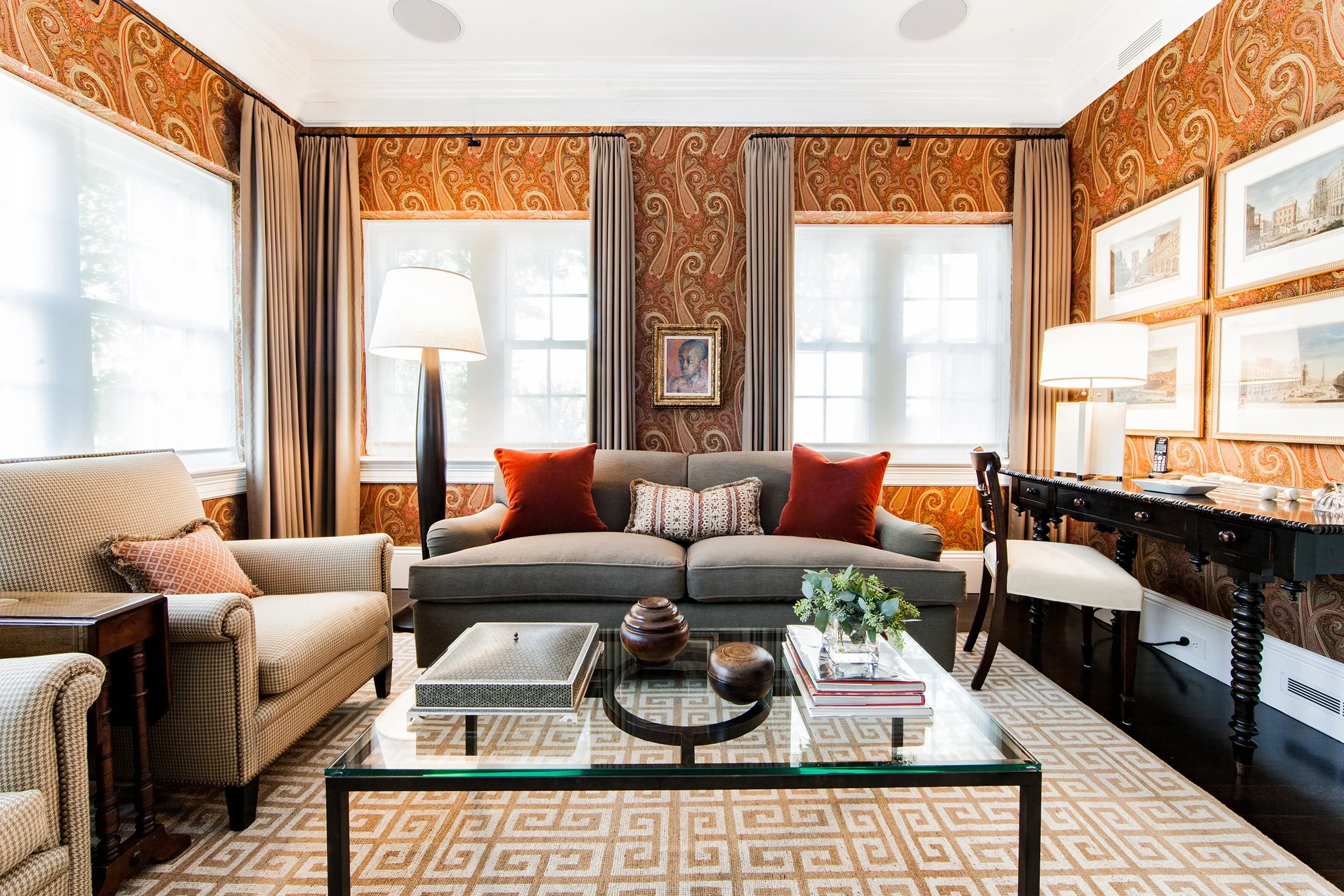 Elegant living room with a gray sofa, orange pillows, armchairs, glass coffee table, patterned rug, artwork, and paisley wallpaper.